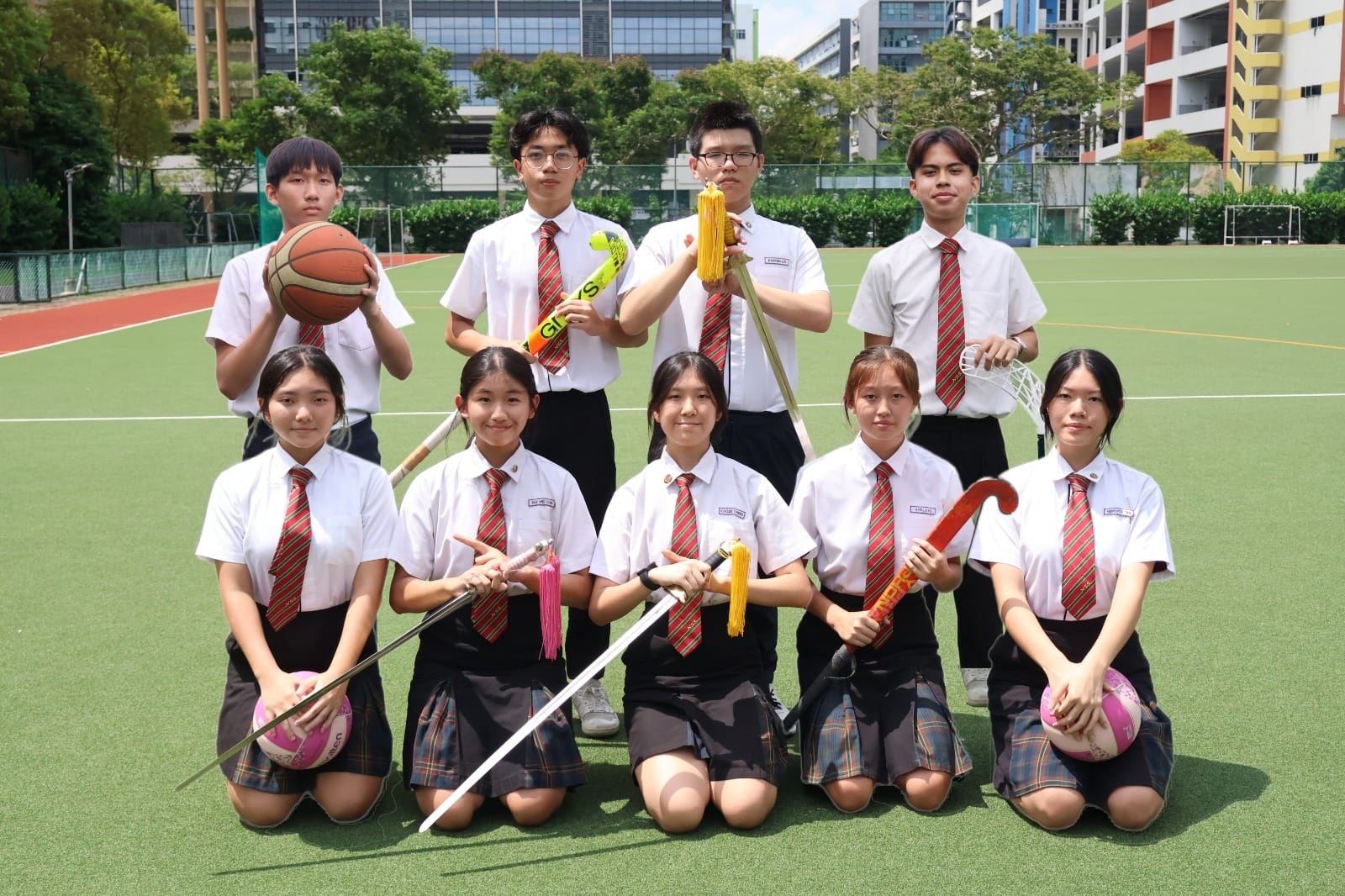 Nine school students in white uniforms with red ties pose on a sports field holding various sports equipment.