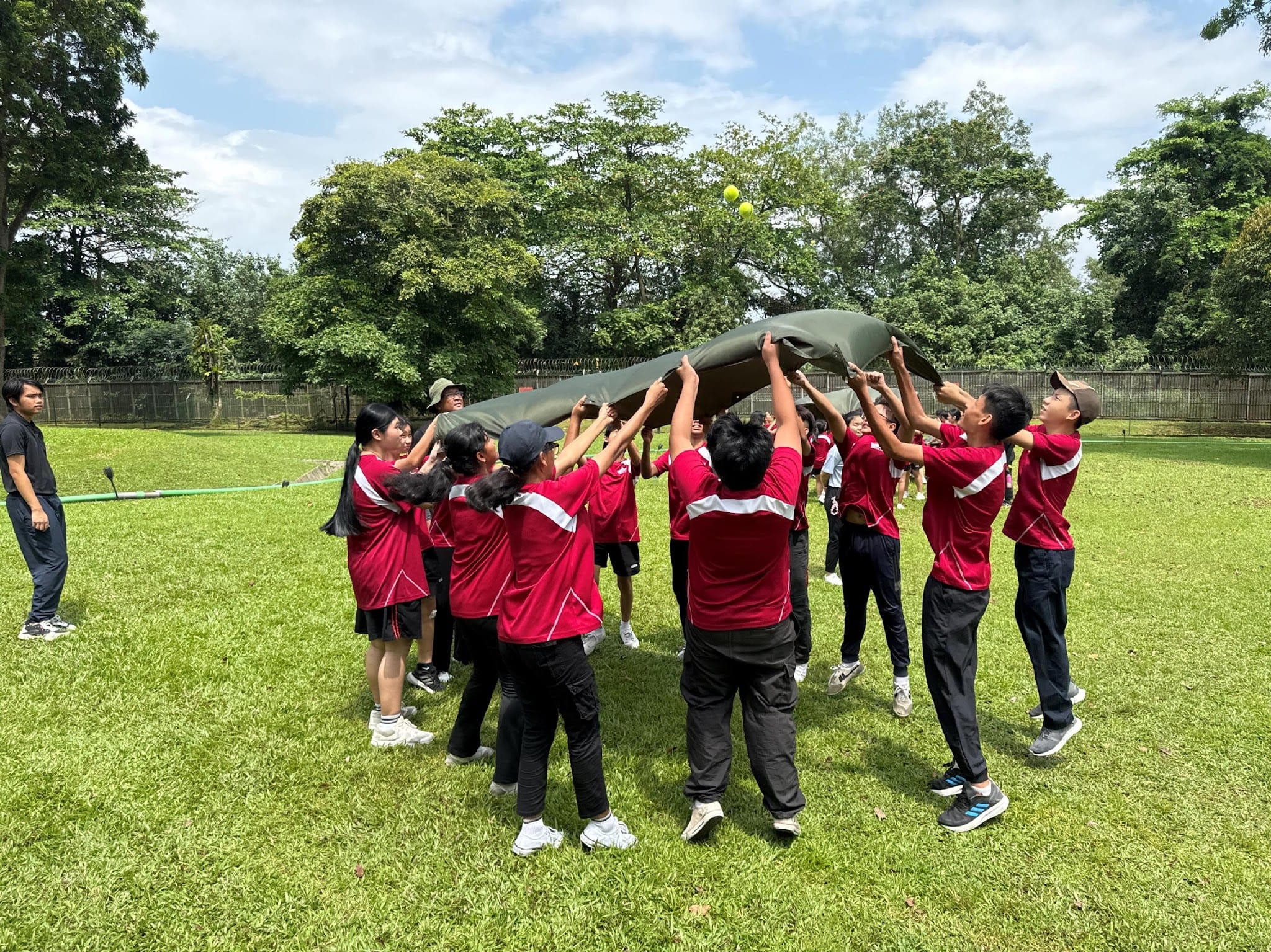 Students in red shirts hold up a green tarp outdoors on grass, tossing a ball into the air as a team activity.