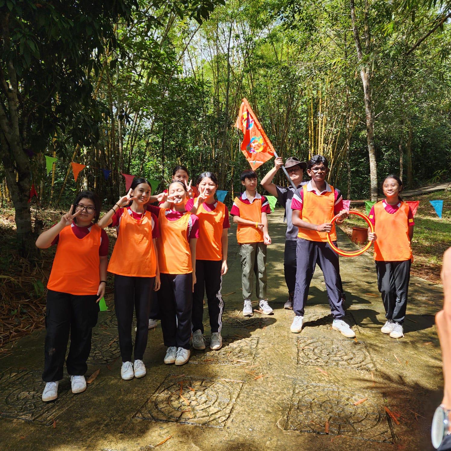 A group of students in orange vests pose outdoors in a bamboo forest, one holding a flag and another an orange hoop.