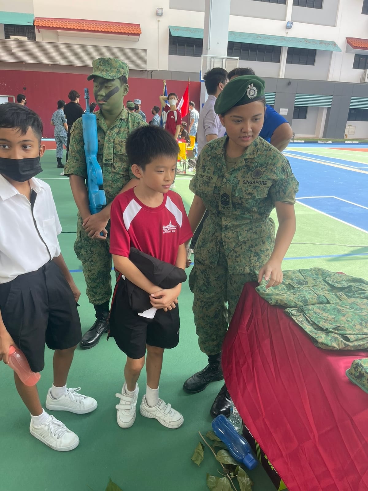 Two Singapore NCC cadets in camouflage uniforms interact with young students at an outdoor school event.