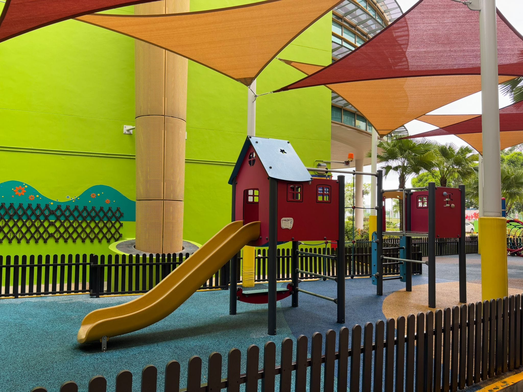Colorful outdoor playground with a red playhouse, yellow slide, black fence, and orange shade sails overhead.