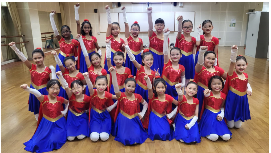 Group of young girls in red sequin and blue dance costumes posing with raised fists in a dance studio.