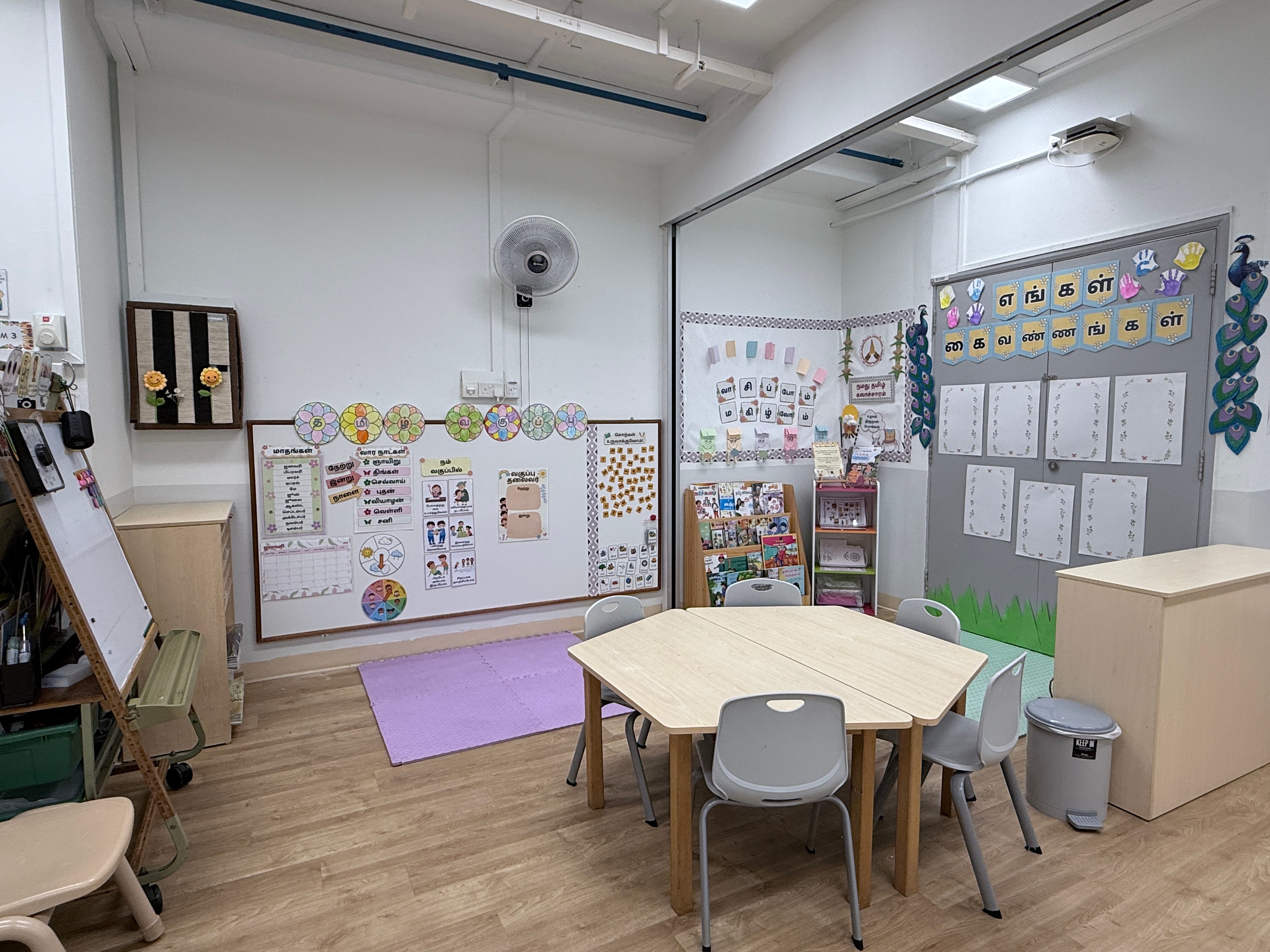 A bright children's classroom with Tamil script displays, a hexagonal table with small chairs, a bookshelf, and colorful wall charts.