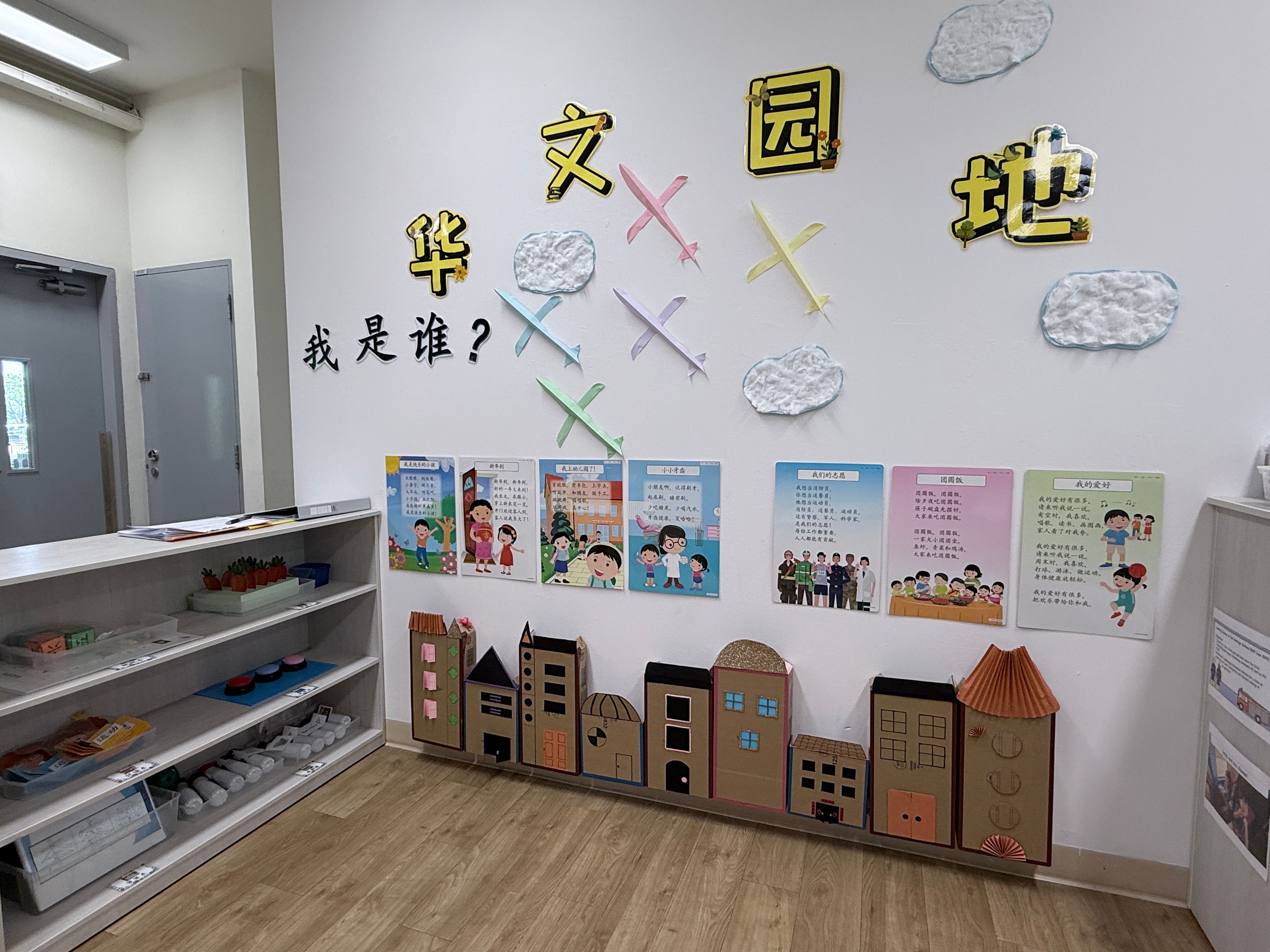 Classroom wall with large Chinese characters, colorful paper airplanes, cloud decorations, children's posters, and cardboard building cutouts below.