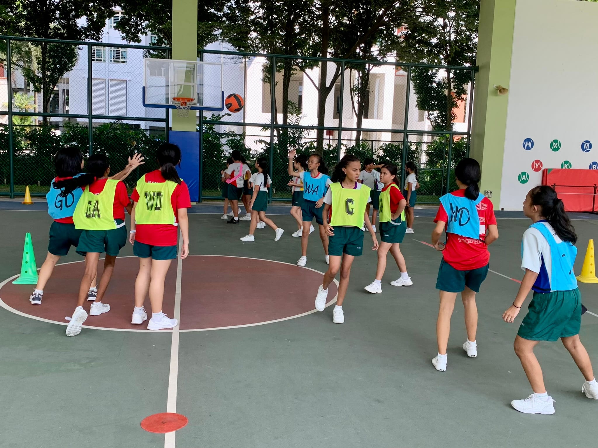 Girls in colored position bibs play netball on an outdoor court with a ball in the air near the hoop.