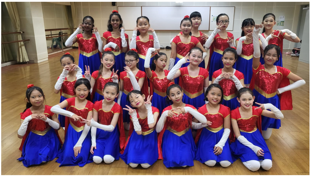 A group of young girls in red sequined tops and blue skirts with white gloves posing in a dance studio.