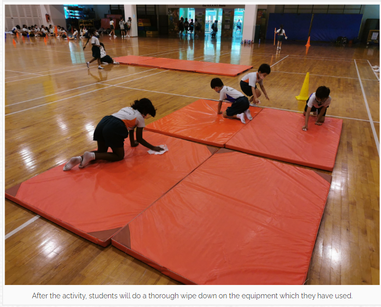 Three children in uniforms kneel on orange gymnastics mats in a school gym, wiping them clean.
