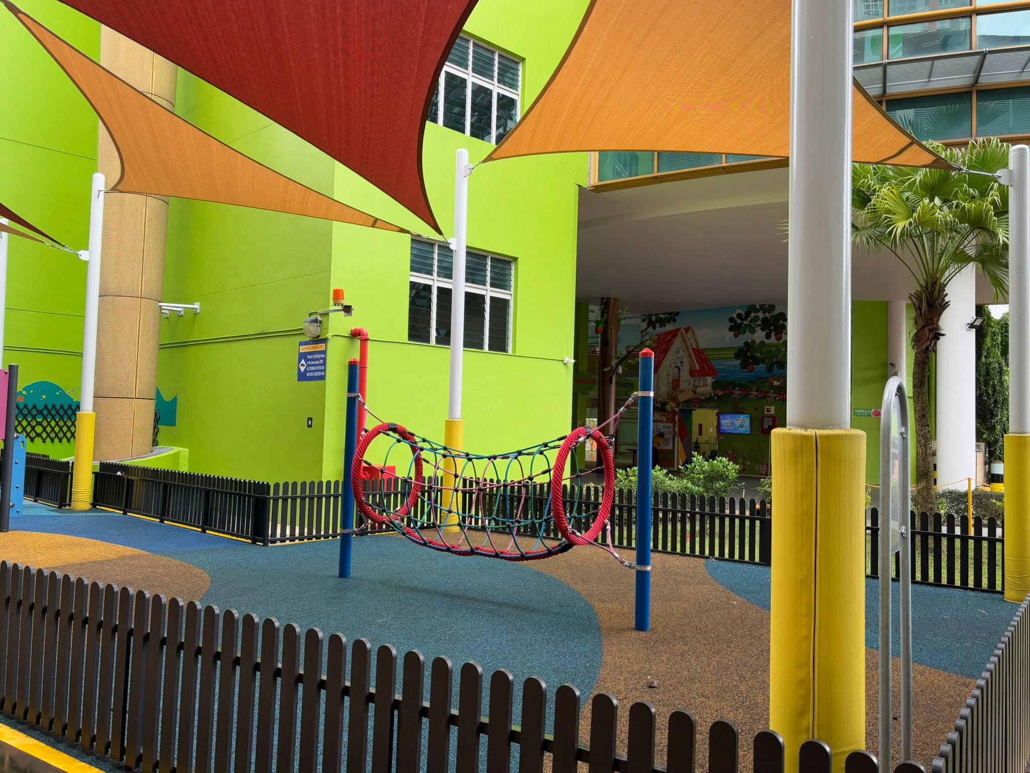 Colorful children's playground with rope tunnel, shade sails, rubber flooring, and bright green building walls.