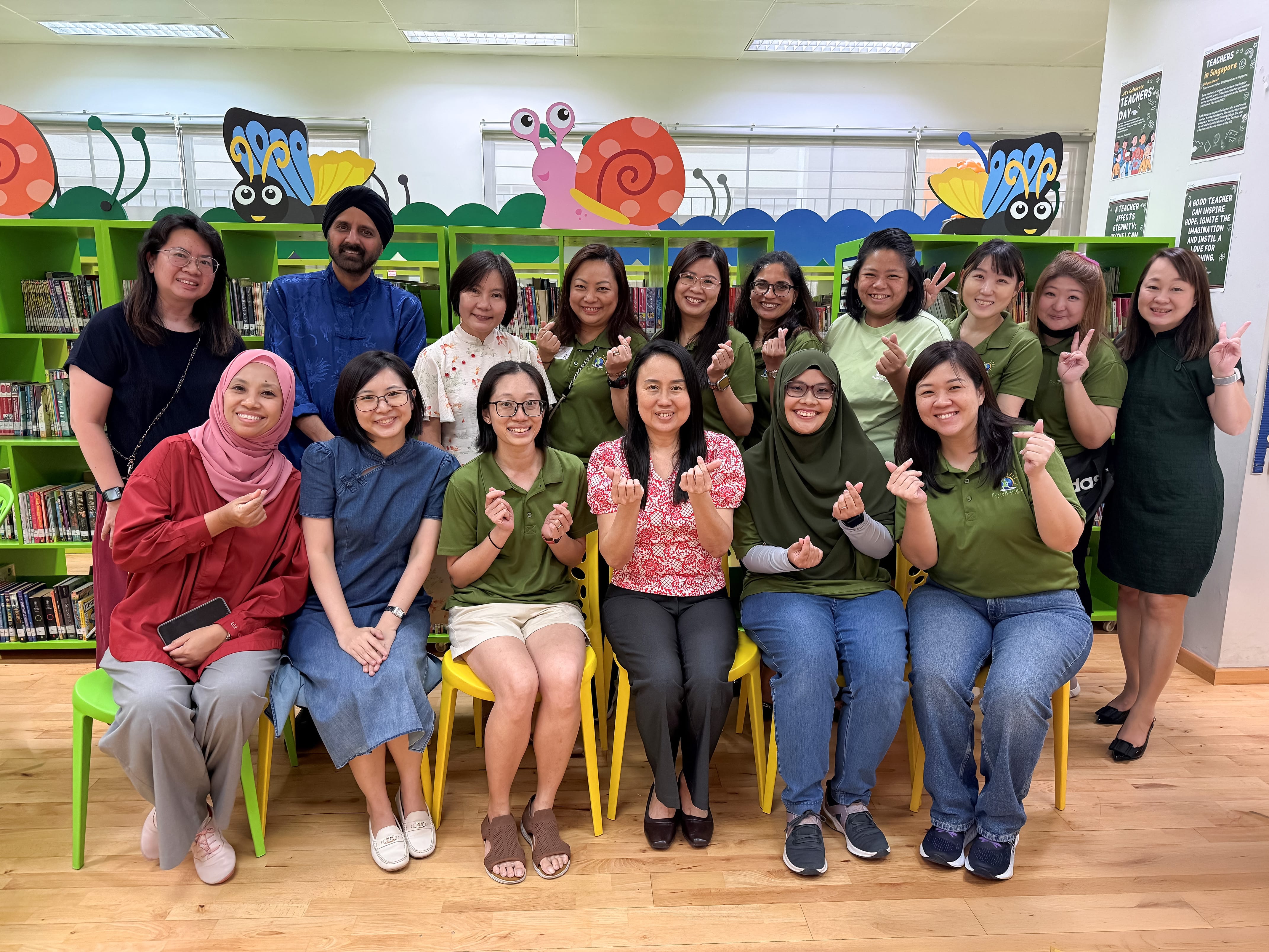 A group of about 15 smiling adults pose in a colorful school library with green shelves and cartoon decorations.