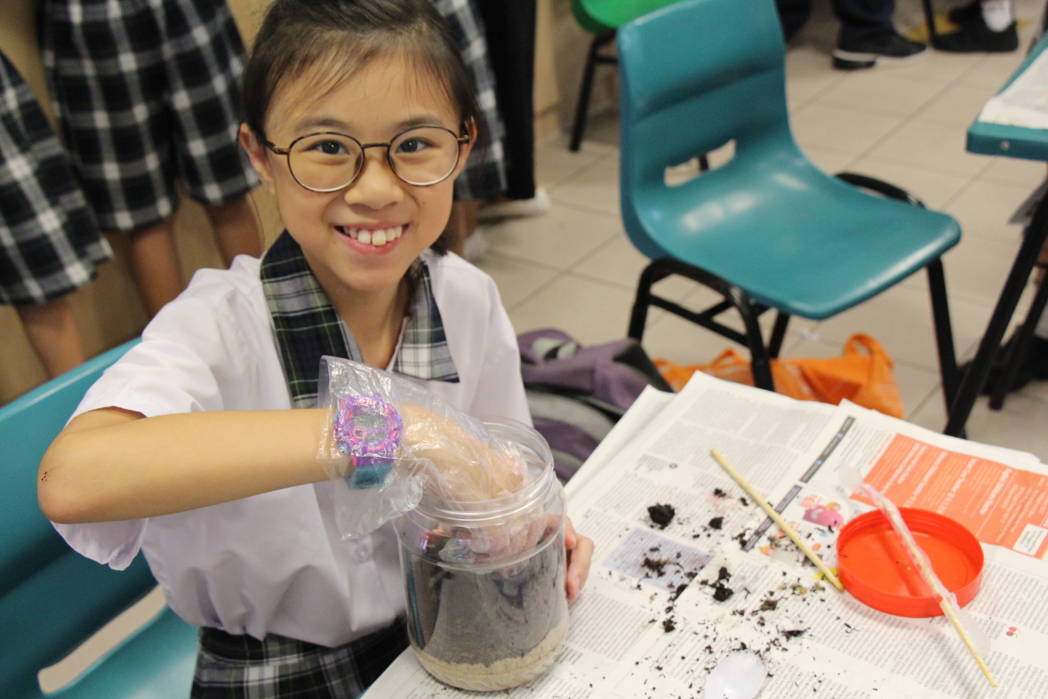 Student holds a plastic jar with soil at a desk covered in dirt and a chopstick.