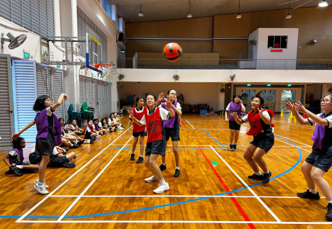 Students in red and purple vests play basketball in an indoor gym with a ball mid-air near the hoop.