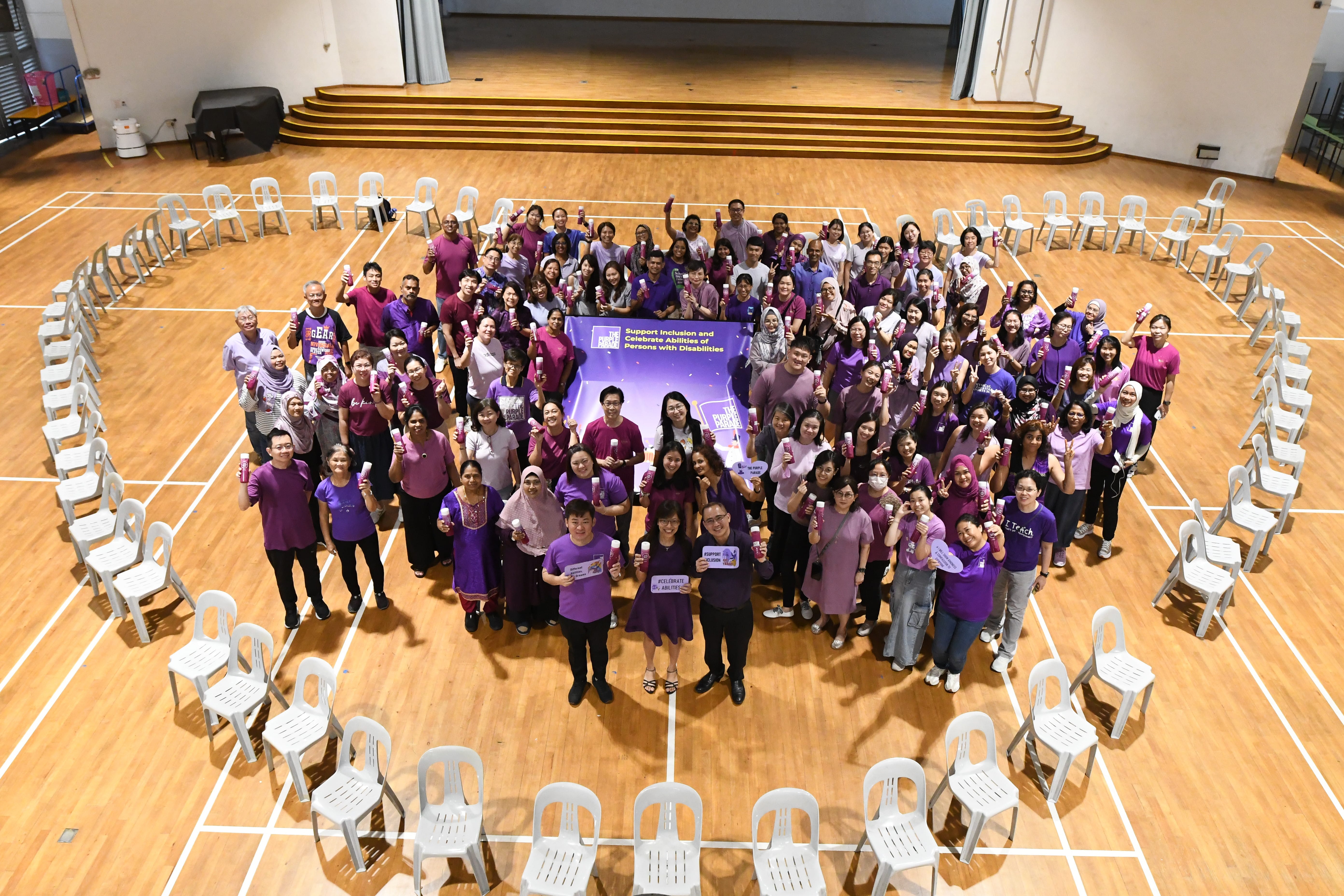 Group in purple shirts holding signs and items on a court, surrounded by chairs arranged in a heart shape.