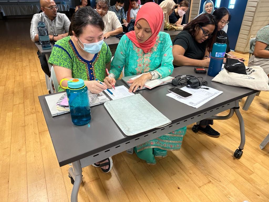 A group of people sit at tables in a classroom, with two women writing on papers together in the foreground.