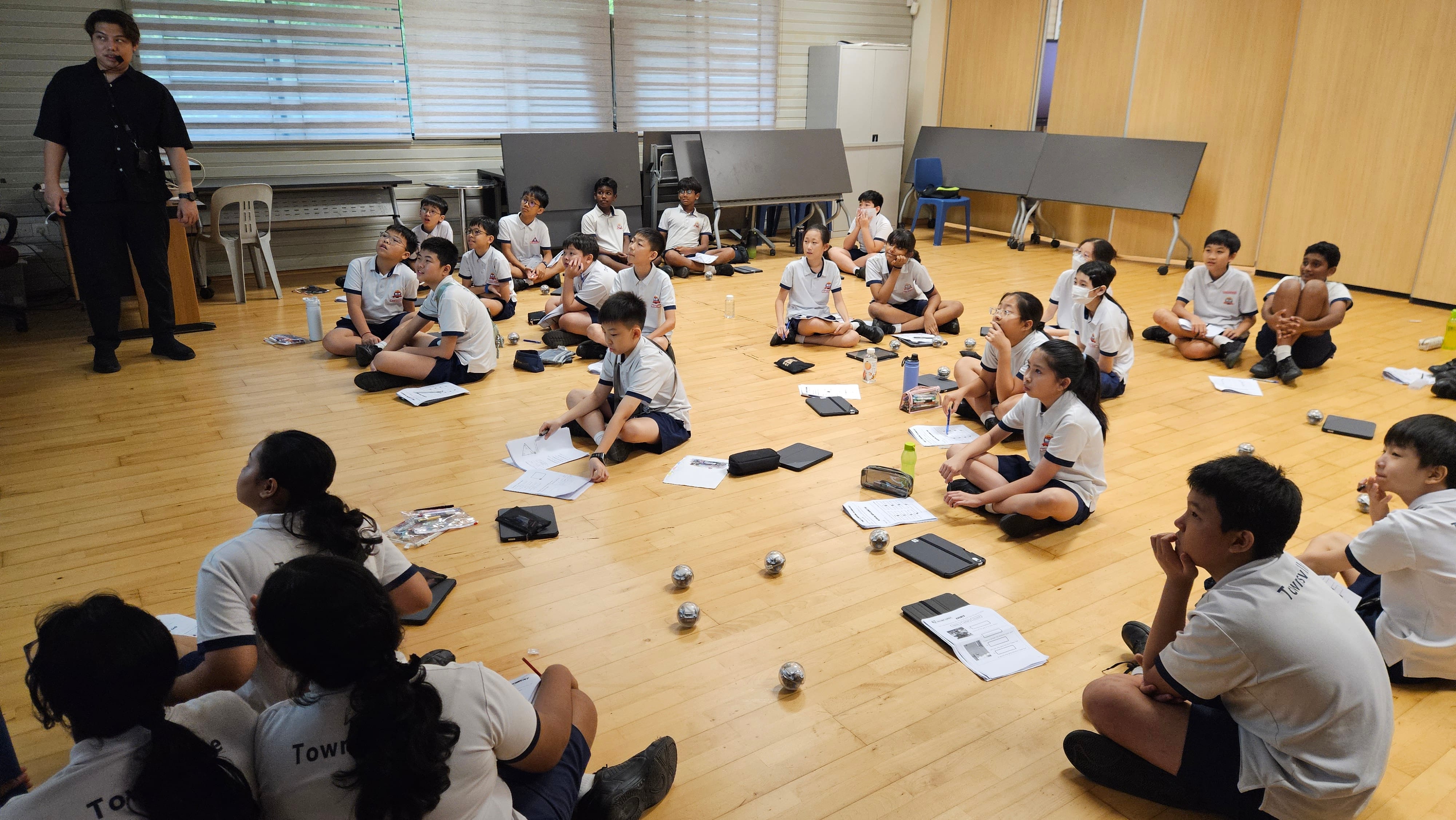 A teacher in black stands while uniformed students sit on a wooden floor with papers and small robots during a lesson.