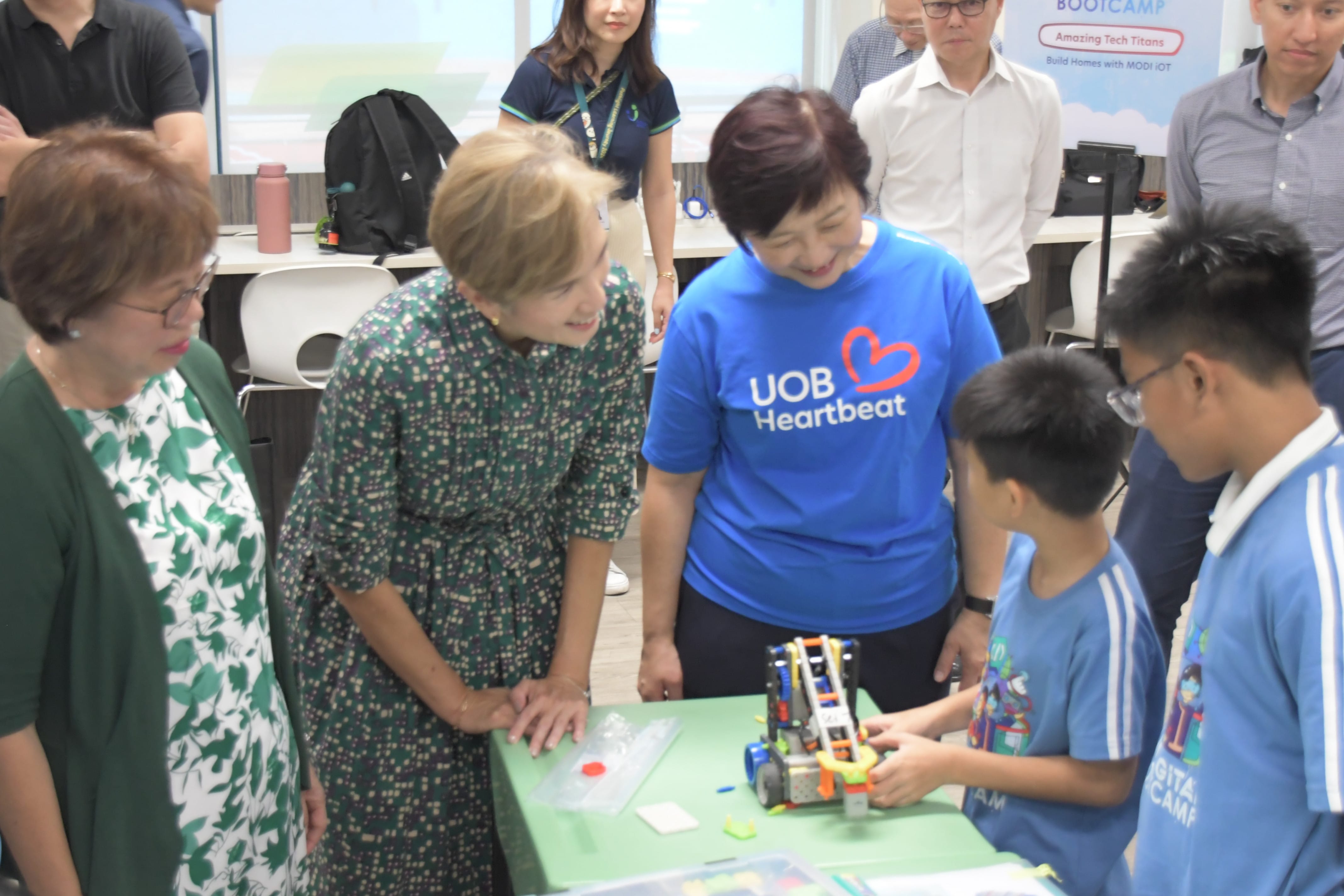 Adults including a UOB Heartbeat volunteer lean over a table watching two boys demonstrate a colorful toy robot.