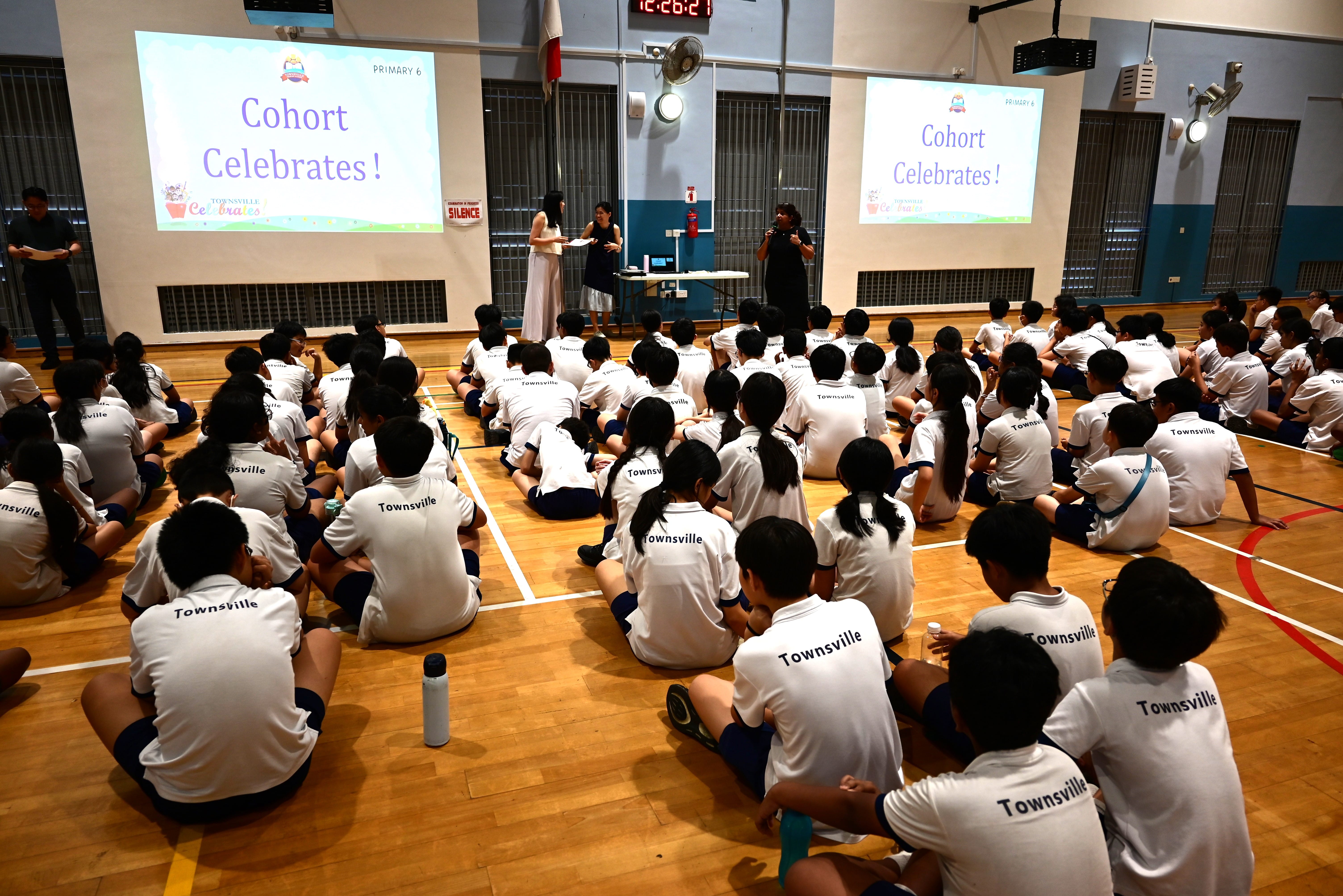 Students seated on a gym floor facing presenters; two screens read Cohort Celebrates!