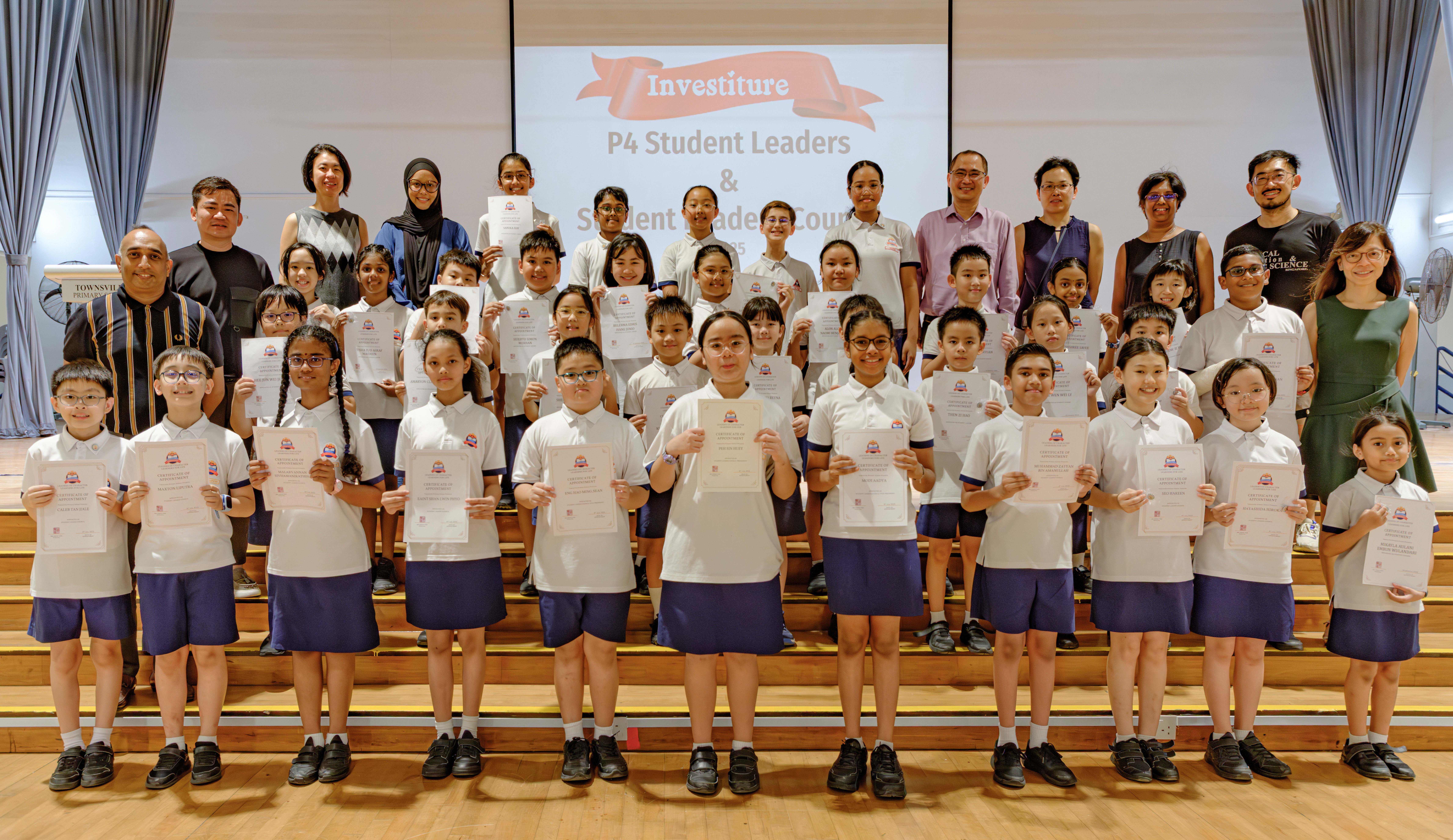 Group of students in school uniforms hold certificates on stage, with teachers behind and Investiture banner.