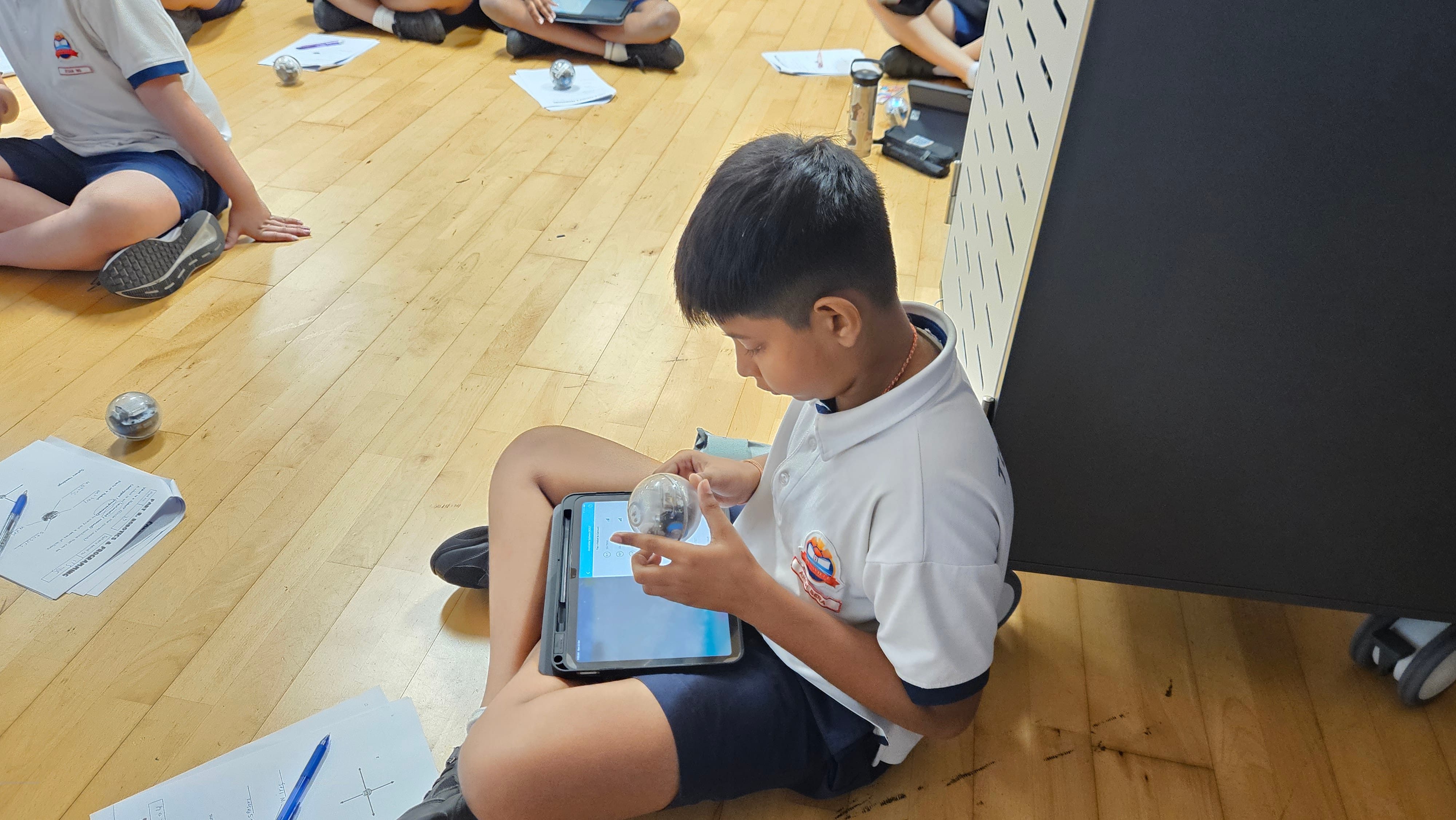 A boy in school uniform sits on a wooden floor, holding a small spherical robot while tapping a tablet screen.