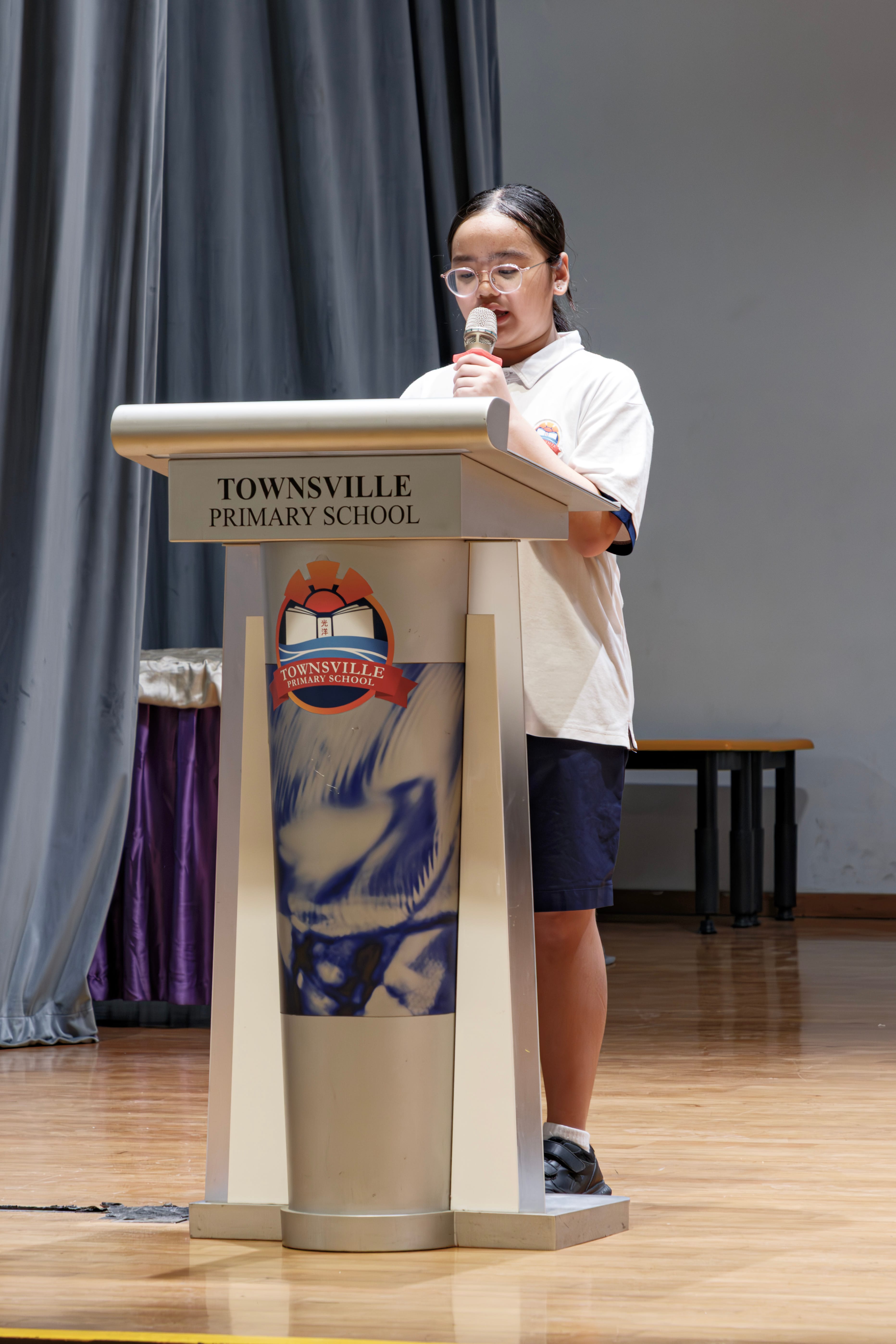 A young girl wearing glasses speaks into a microphone at a Townsville Primary School podium on a wooden stage.