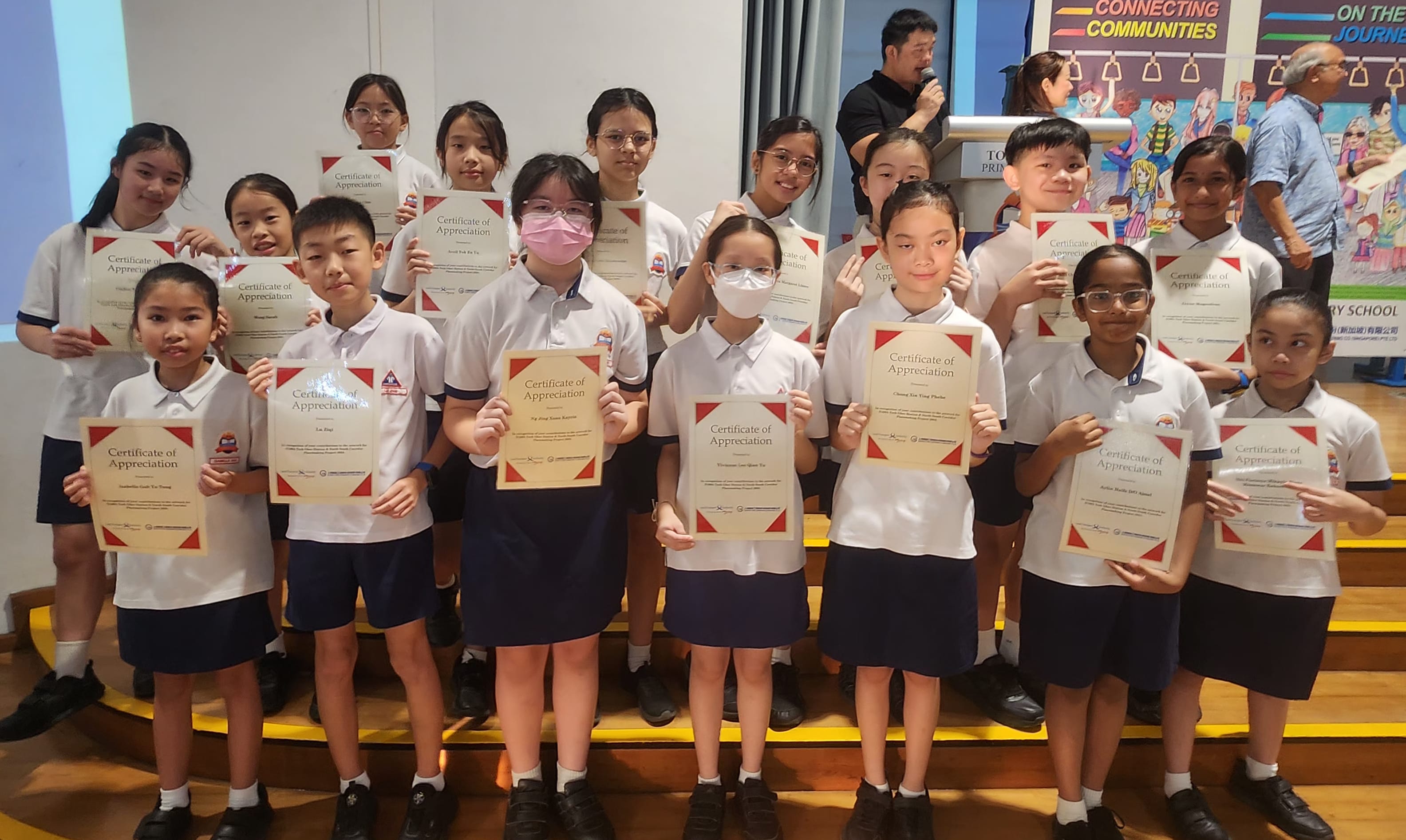 A group of school children in uniforms holding Certificates of Appreciation on a tiered stage.