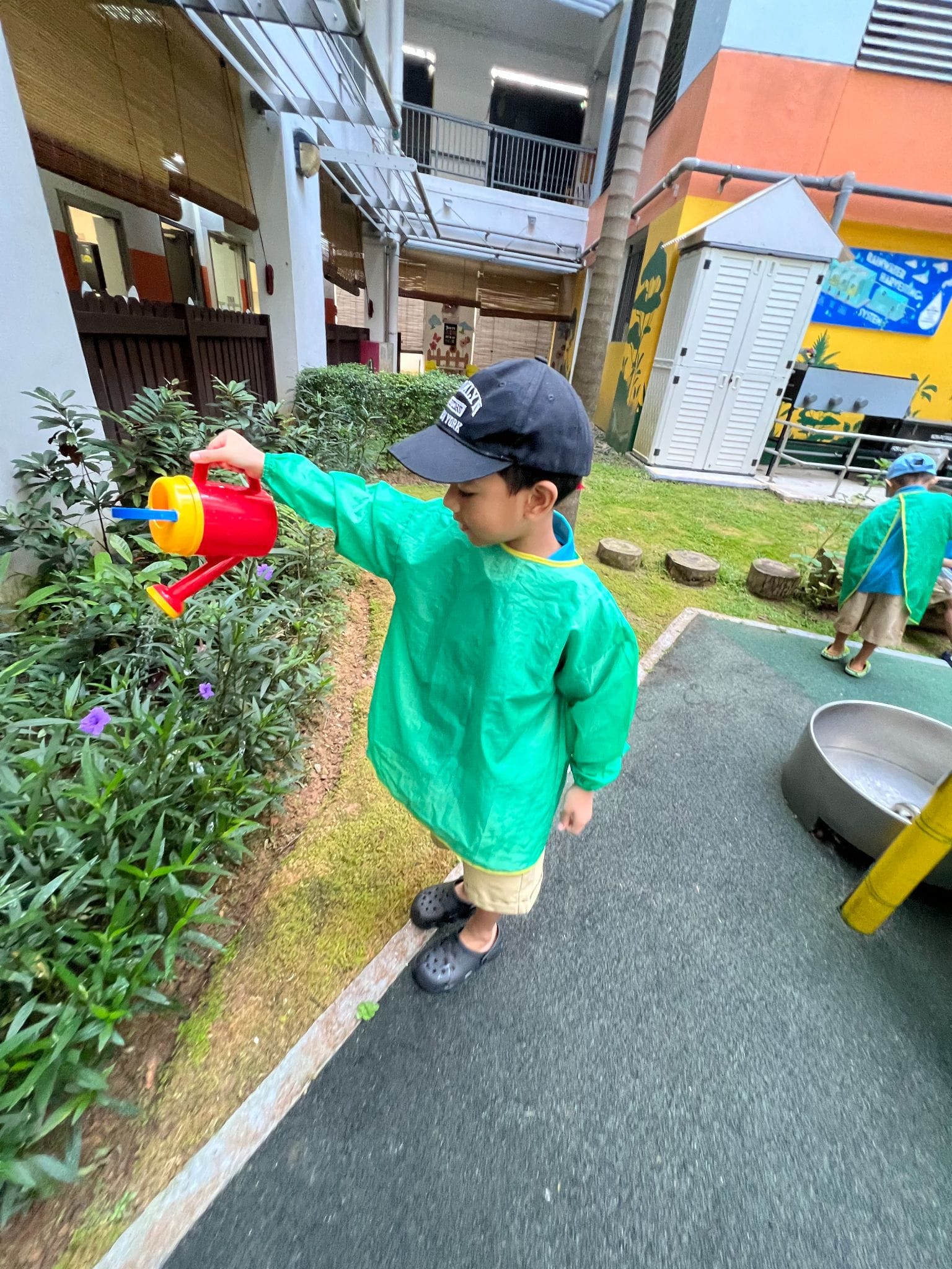 Young child in green smock and cap waters plants with a colorful toy watering can in a school courtyard.