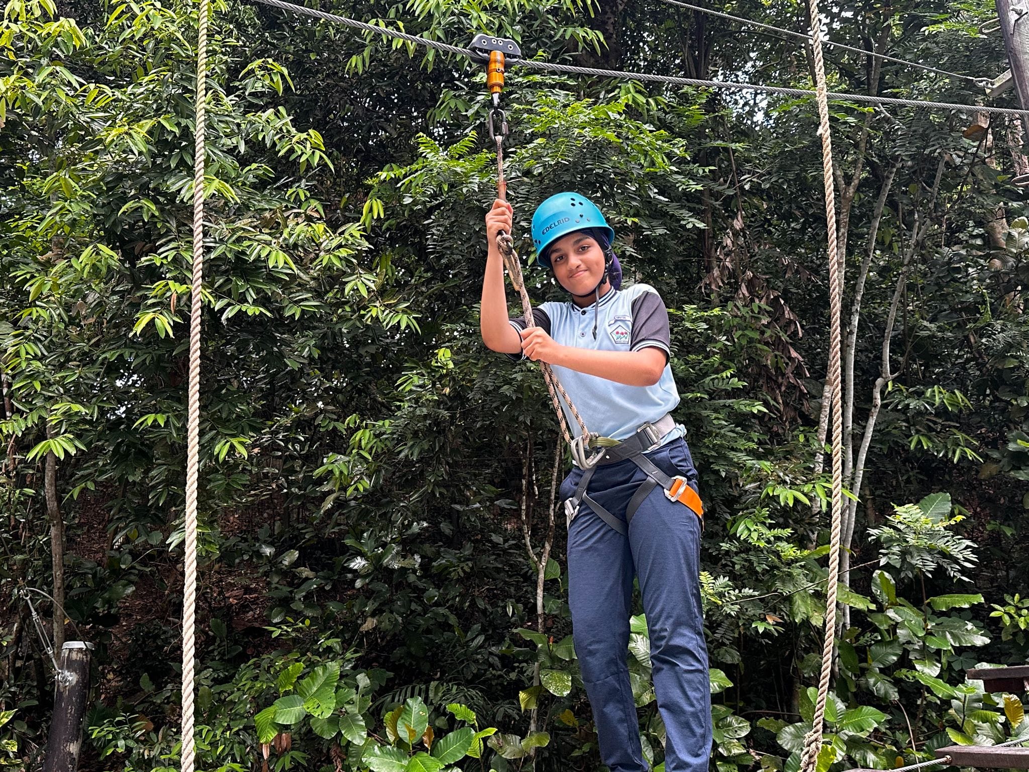 A young person wearing a blue helmet and harness holds a zipline pulley handle surrounded by dense green trees.