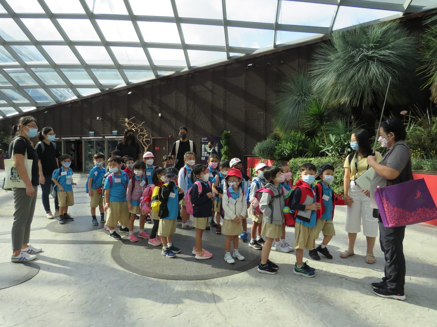 A group of young children in blue uniforms and masks stand with adult chaperones inside a glass-roofed building with plants.