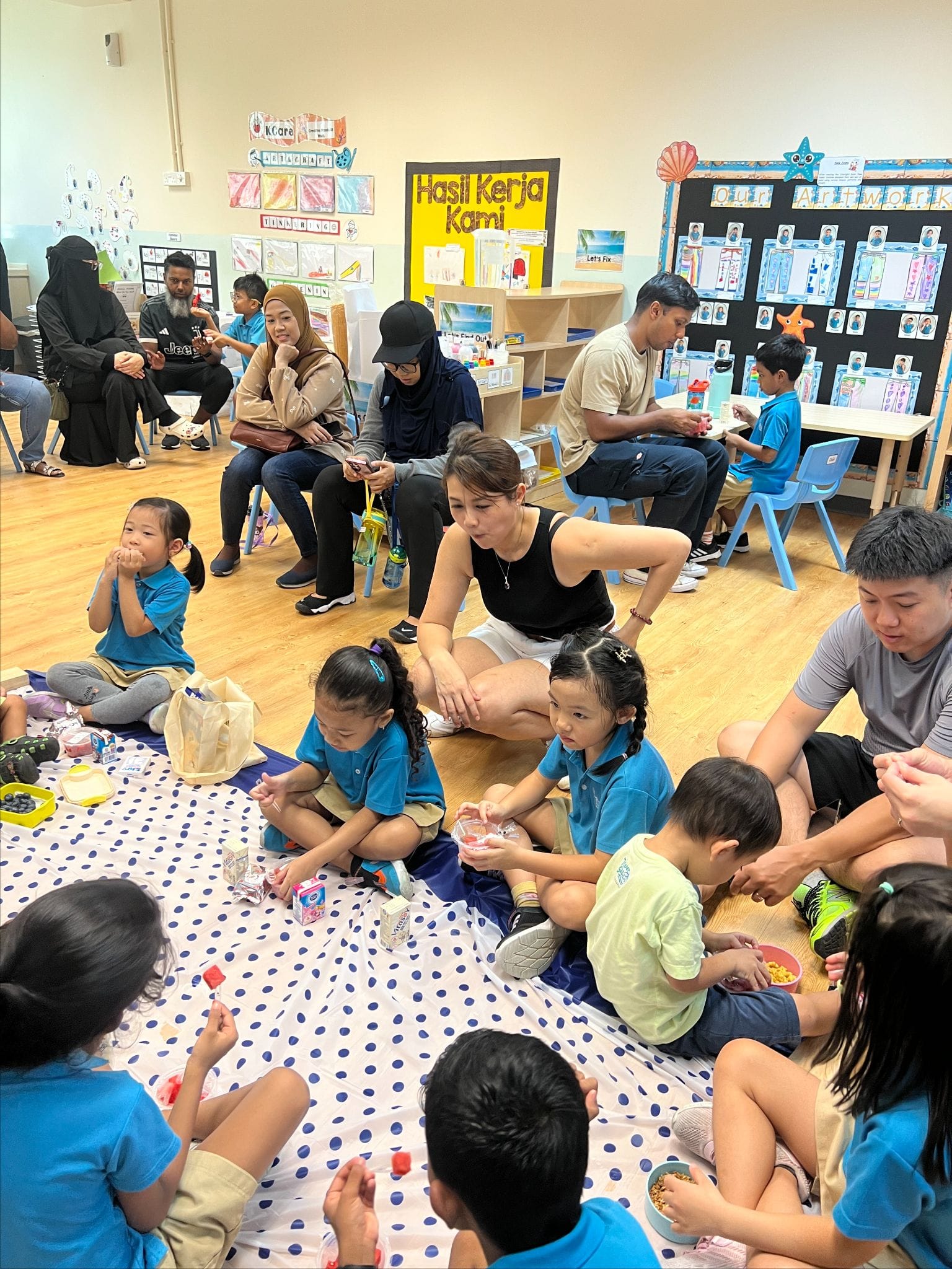 Children in blue uniforms sit on a polka-dot mat eating snacks with adults in a colorful classroom.
