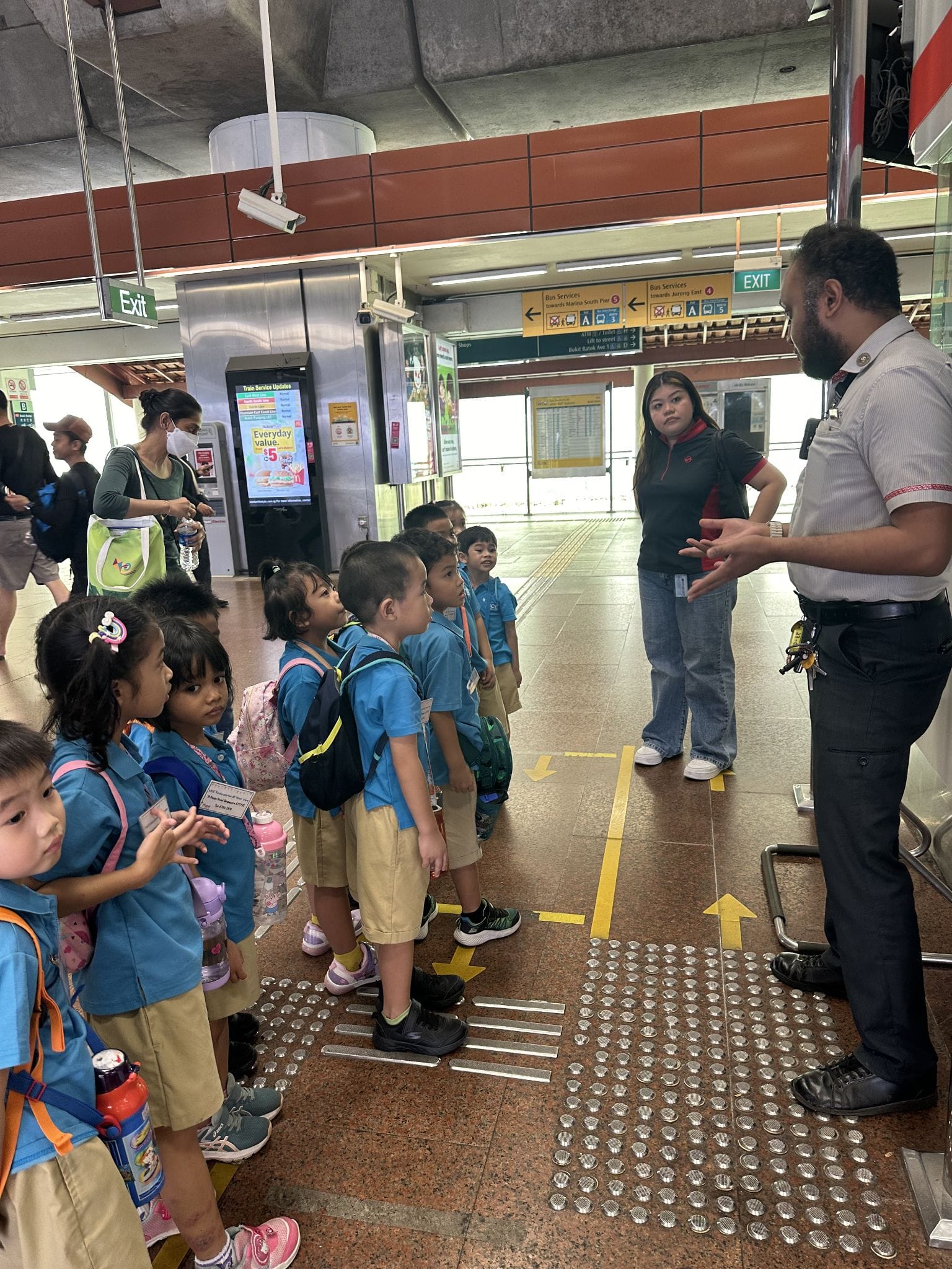A station officer speaks to a group of young schoolchildren in blue uniforms at a Singapore MRT station.