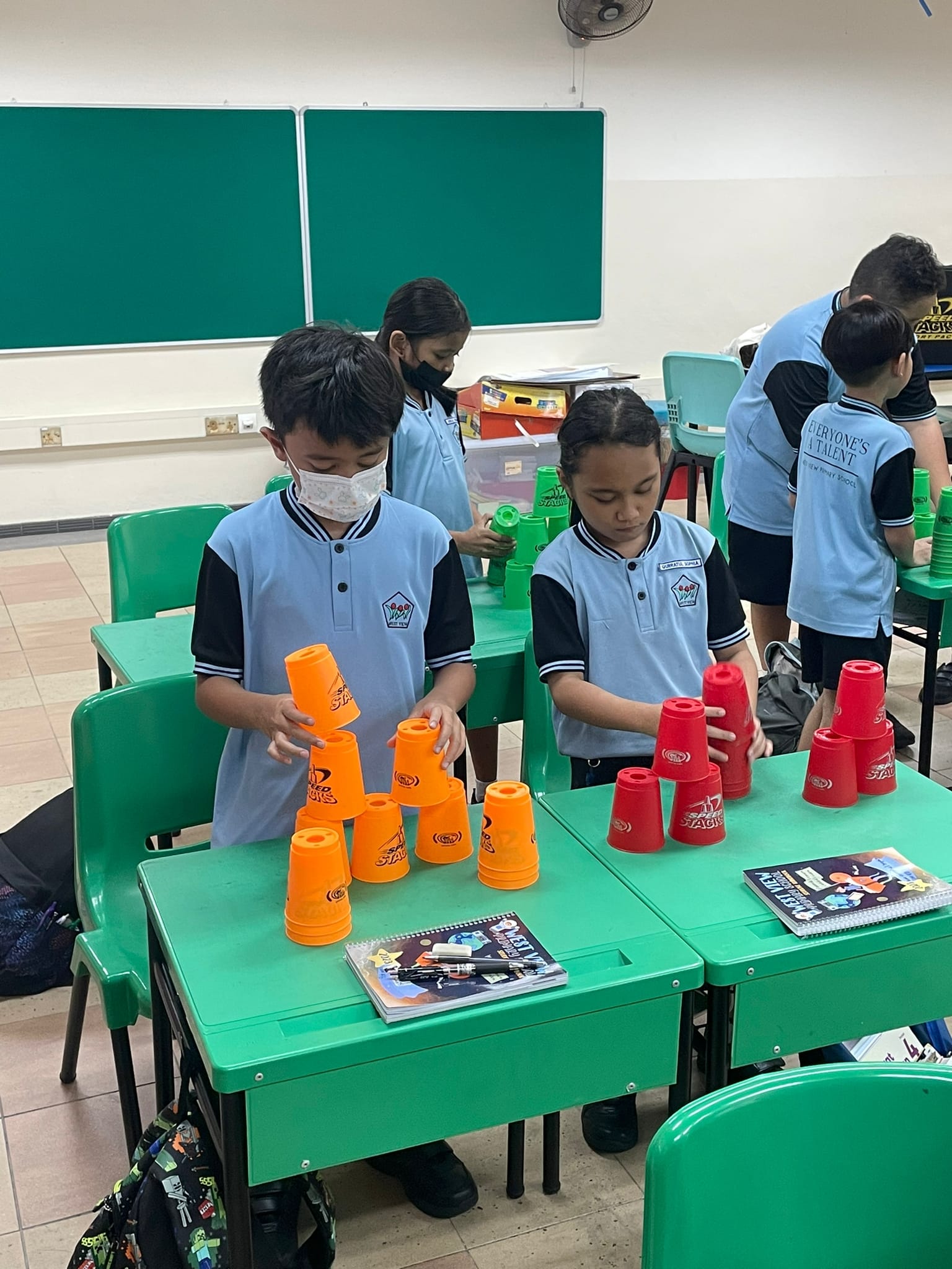 Children in school uniforms stacking orange and red plastic cups on green desks in a classroom.