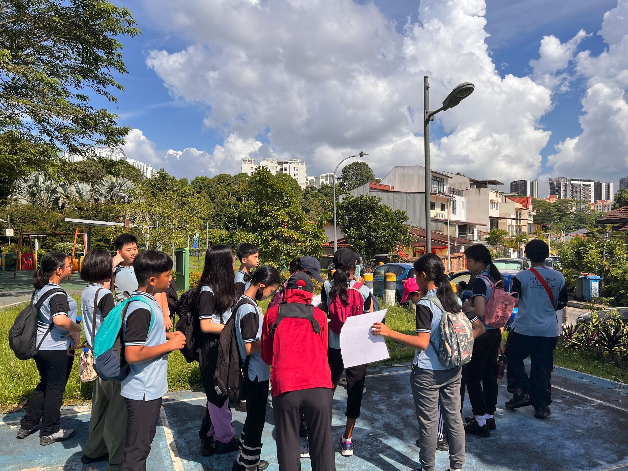 A group of students in school uniforms with backpacks gather outdoors near a playground, one holding a white paper sheet.