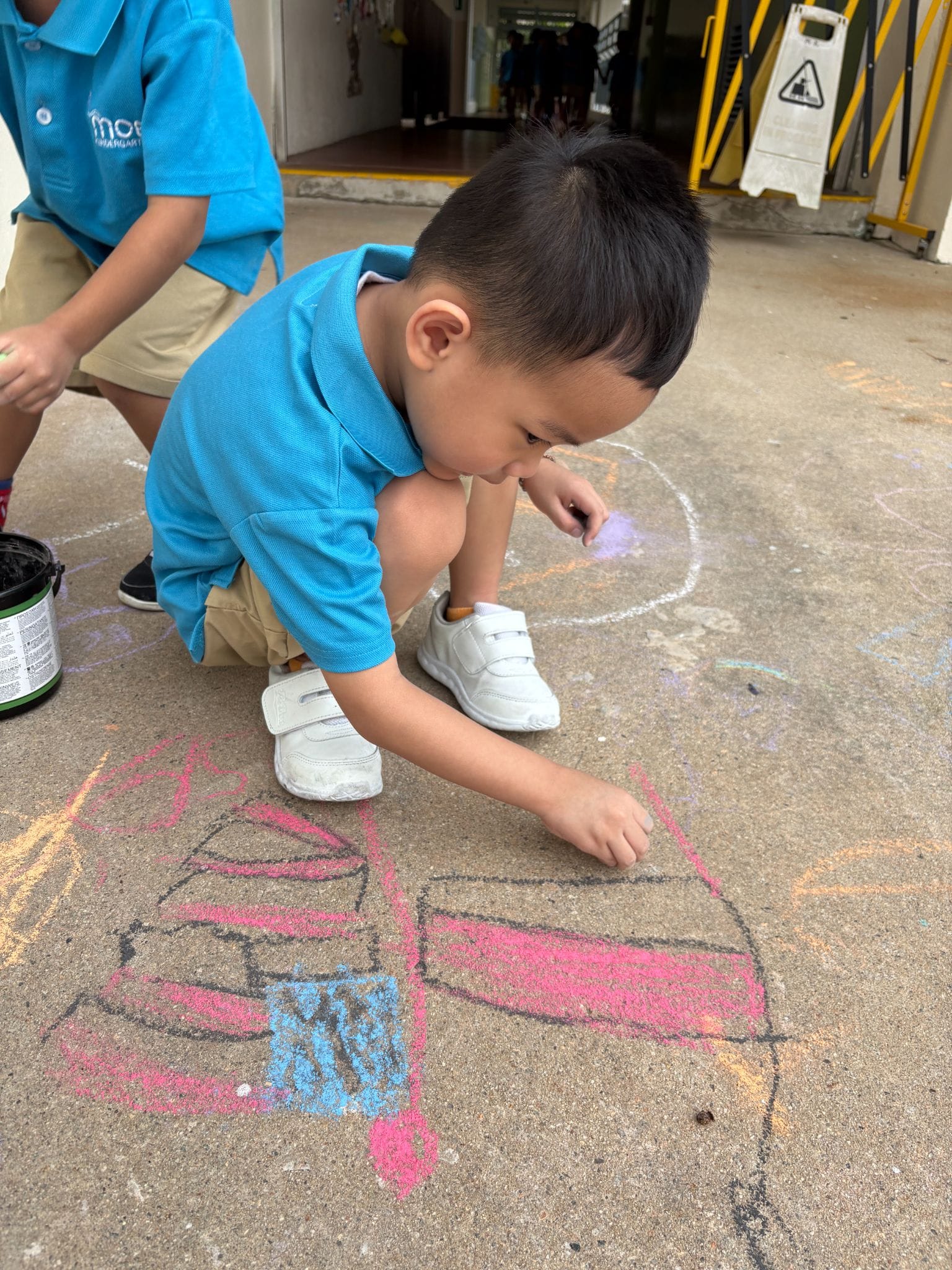 A young boy in a blue shirt crouches on pavement, drawing colorful chalk art on the ground.