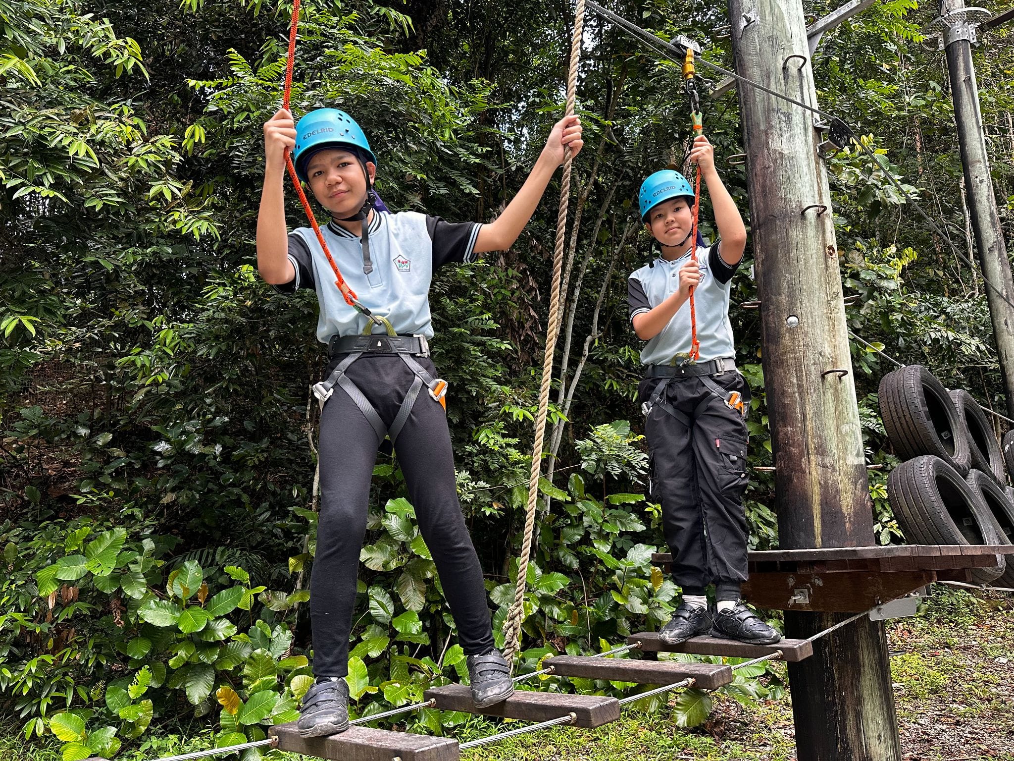 Two children in blue helmets and harnesses stand on a wooden rope course platform surrounded by green trees.