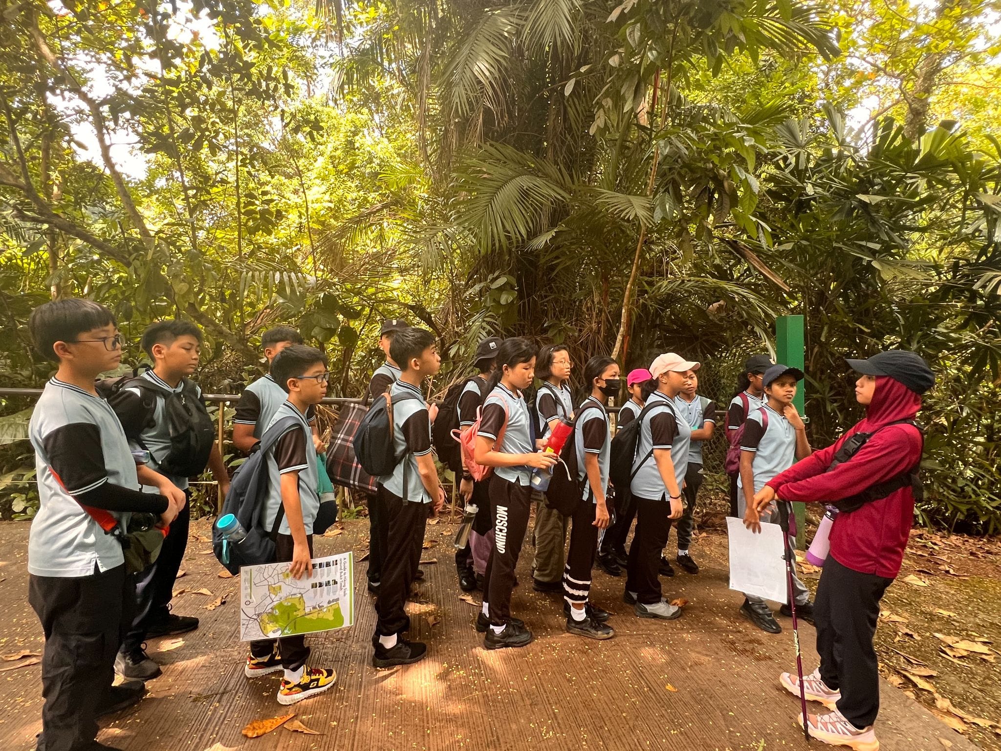 A group of uniformed students with backpacks listens to a guide on a forest trail surrounded by lush trees.