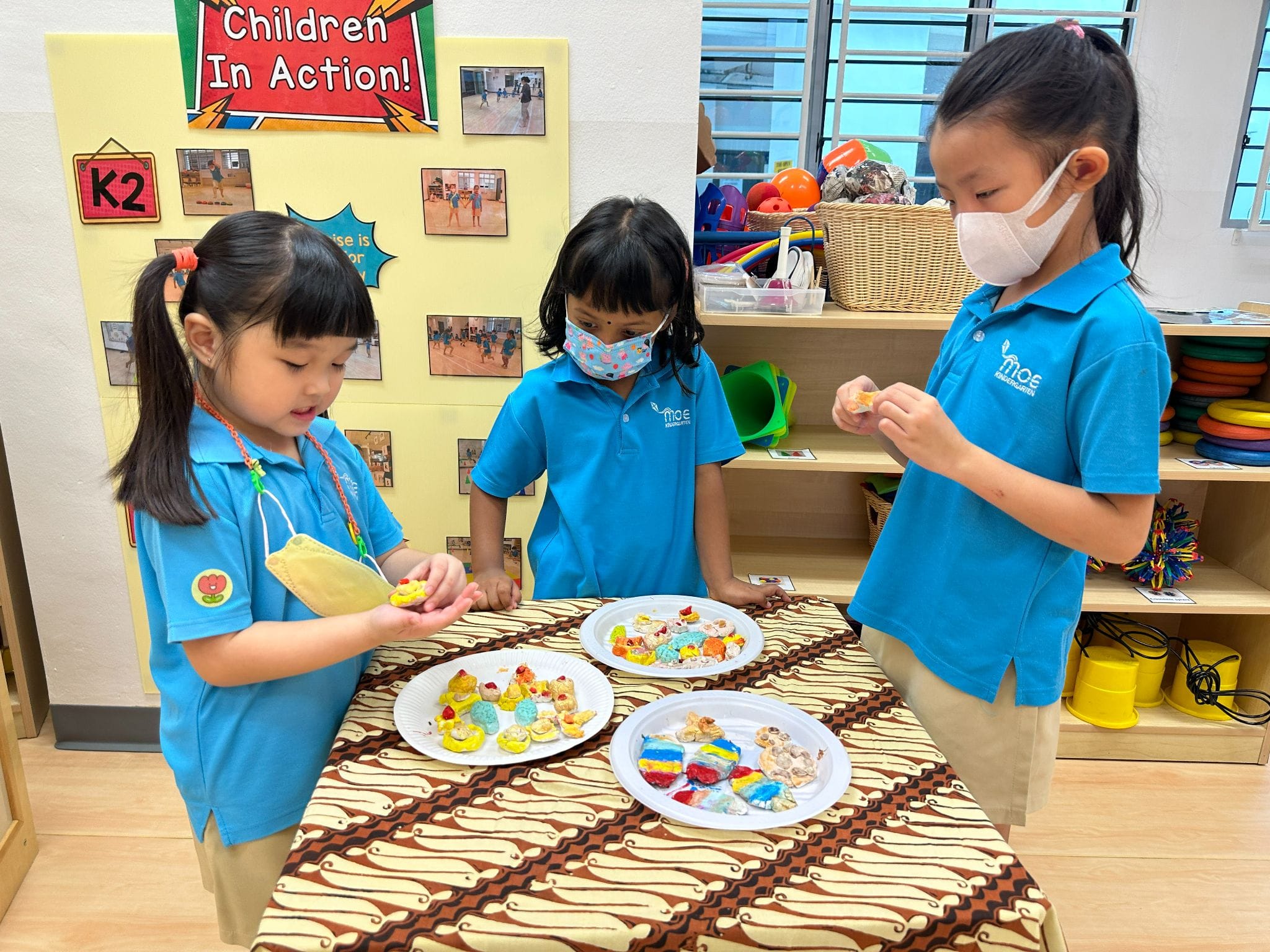 Three young students in blue uniforms stand around a table with plates of colorful decorated cookies in a classroom.