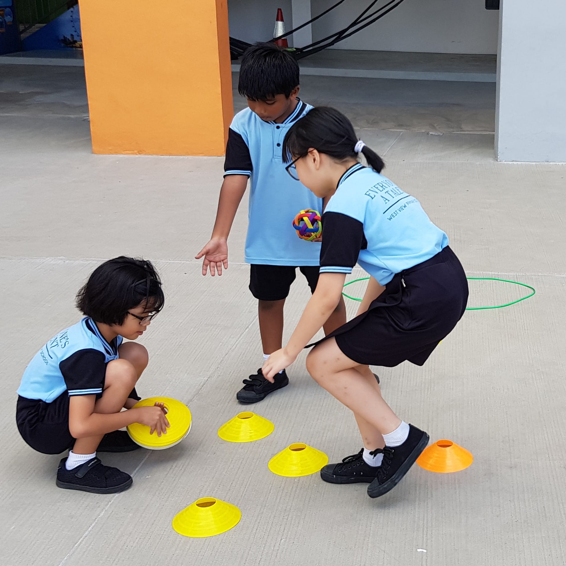Three children in blue school uniforms play outdoors with yellow and orange sport cones and a colorful ball.