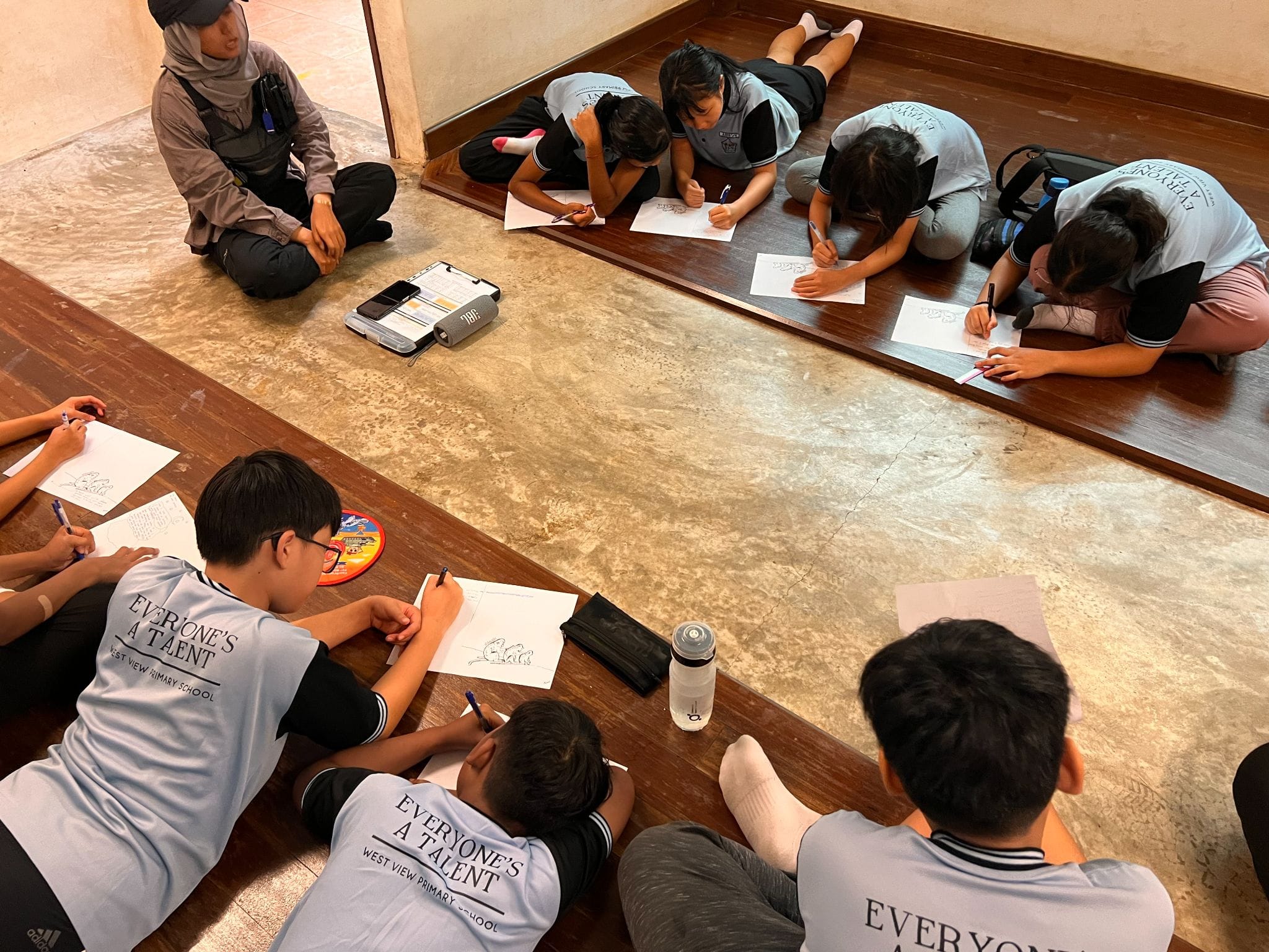 Students in matching shirts reading "Everyone's A Talent" sit and lie on wooden floors drawing on paper.