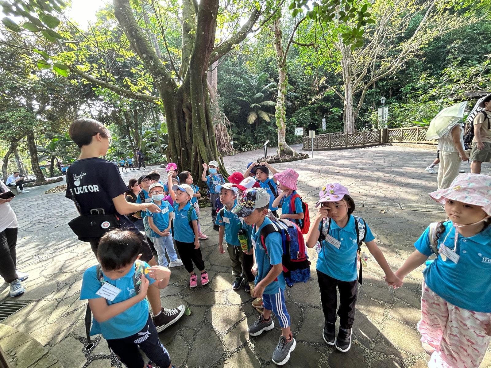 A group of young children in blue uniforms gather around an adult on a paved path surrounded by large trees.
