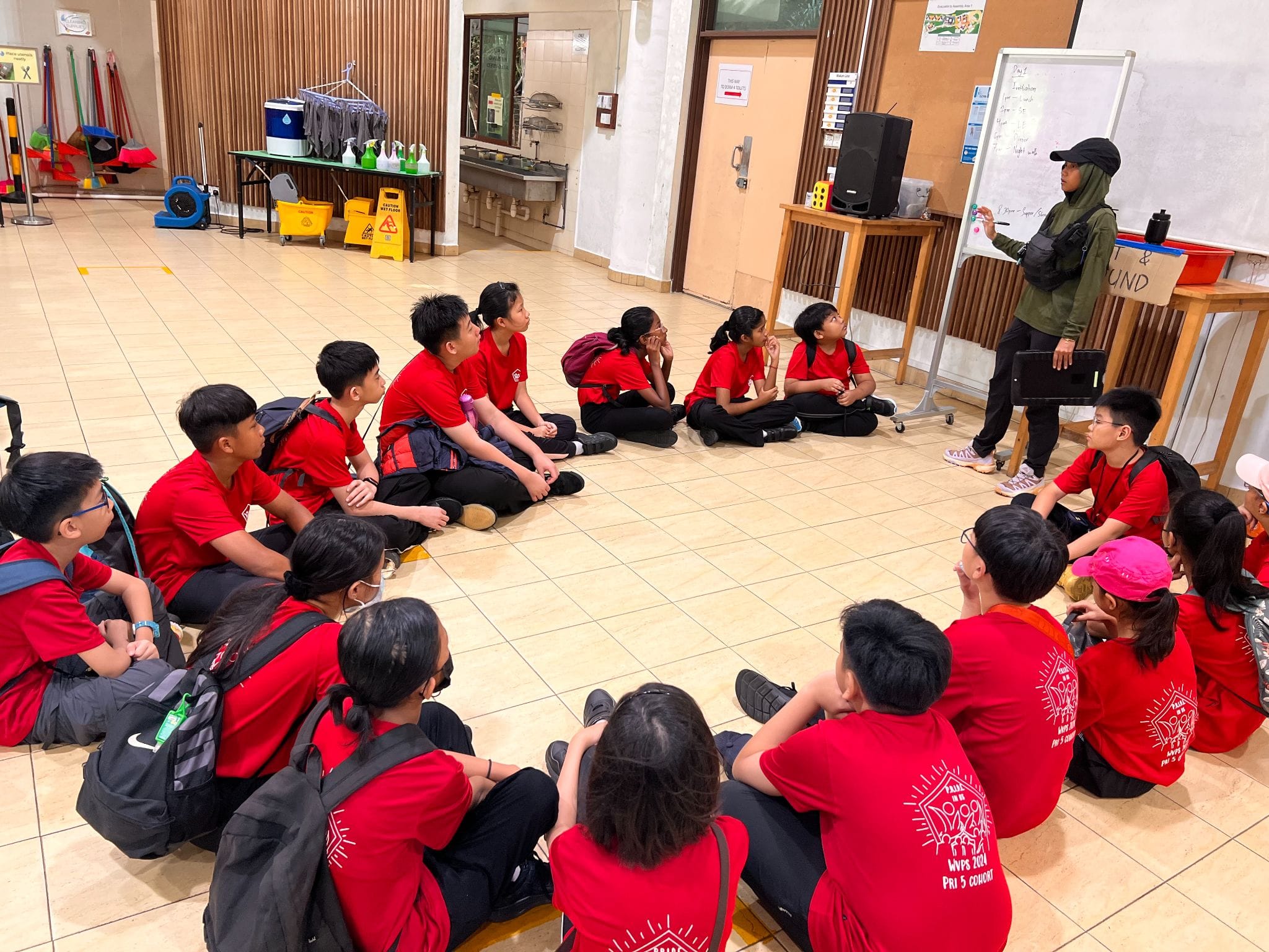 A group of students in red shirts sit on a floor listening to an instructor writing on a whiteboard.