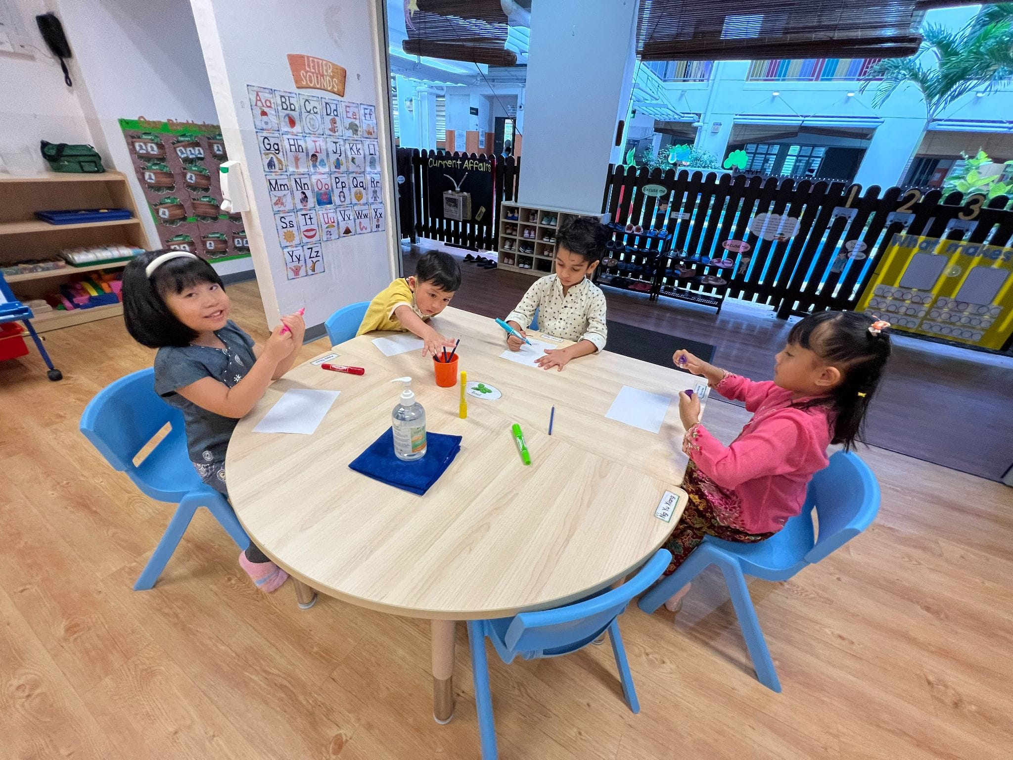 Four young children sit around a round wooden table drawing with markers in a colorful classroom.