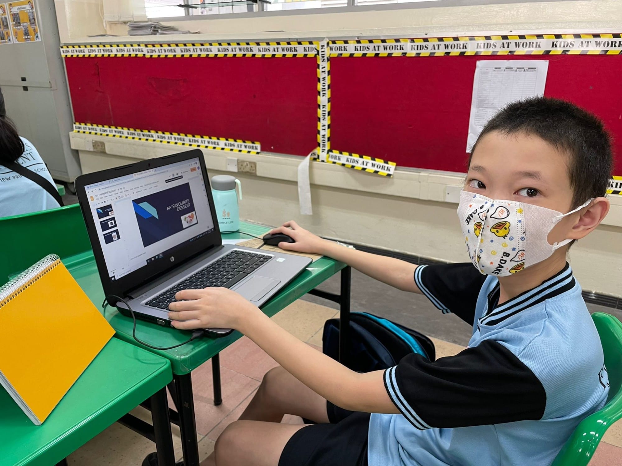 A masked young student in school uniform sits at a green desk working on an HP laptop in a classroom.
