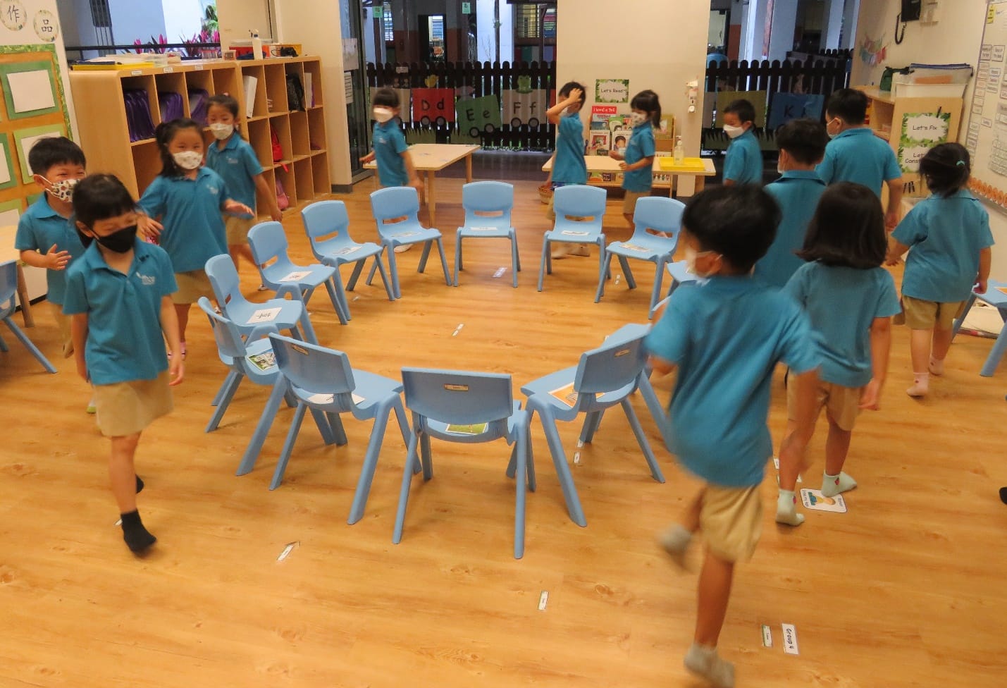 Young masked children in teal uniforms play musical chairs around a circle of blue chairs in a classroom.