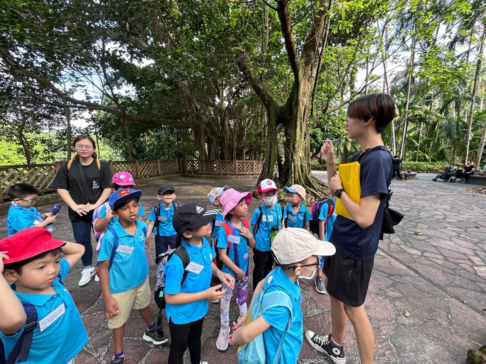 A group of young children in blue uniforms with backpacks listen to a teen guide in a lush park setting.