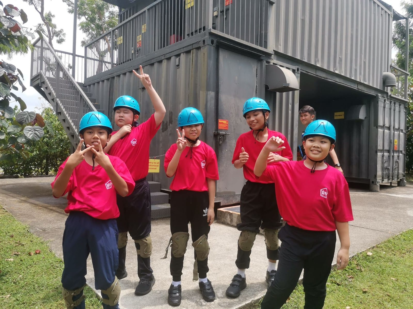 Five children in pink shirts and blue helmets pose with hand gestures in front of a gray metal container structure.