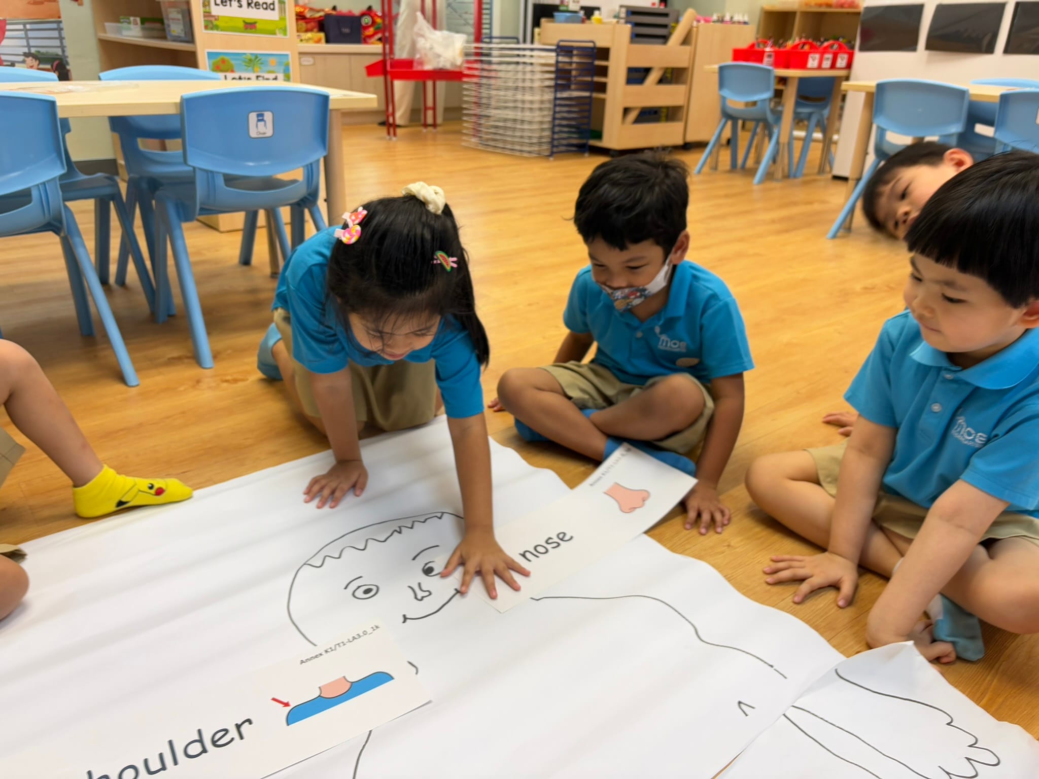 Three young children in blue uniforms sit on a classroom floor working on a large body parts diagram labeled nose and shoulder.