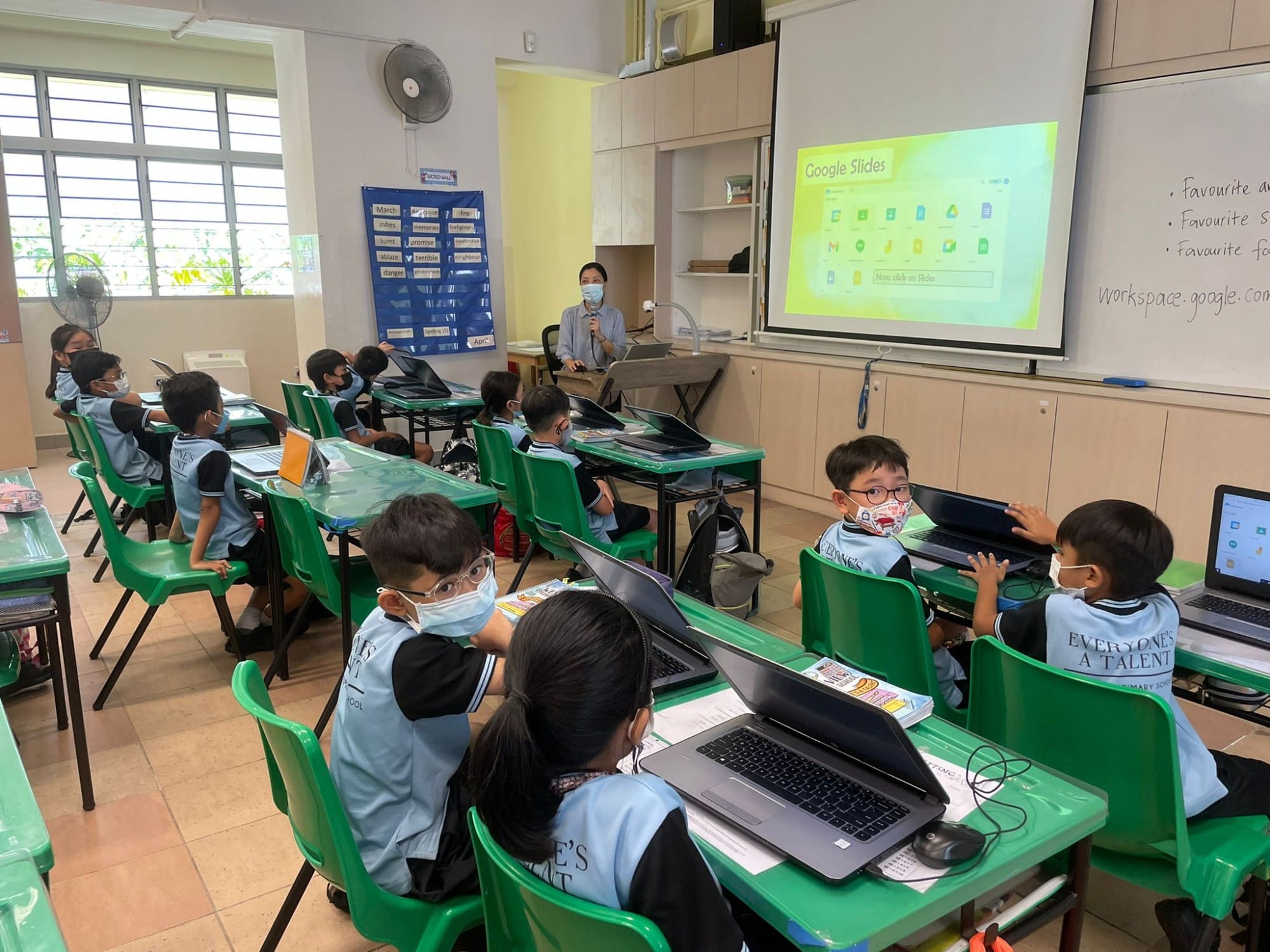 Students in masks sit at green desks with laptops while a masked teacher presents Google Slides on a projector screen.