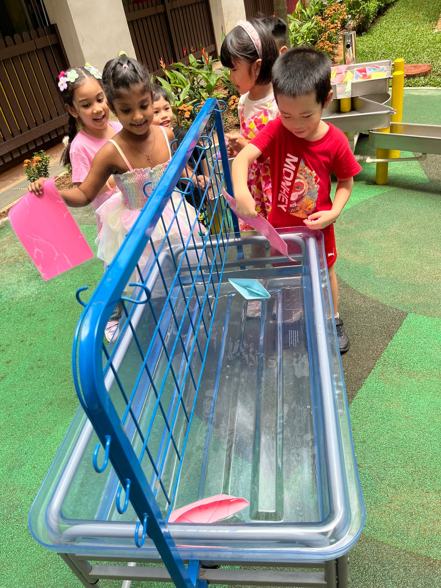 Five young children play with paper boats in a shallow water table outdoors on a green padded ground.