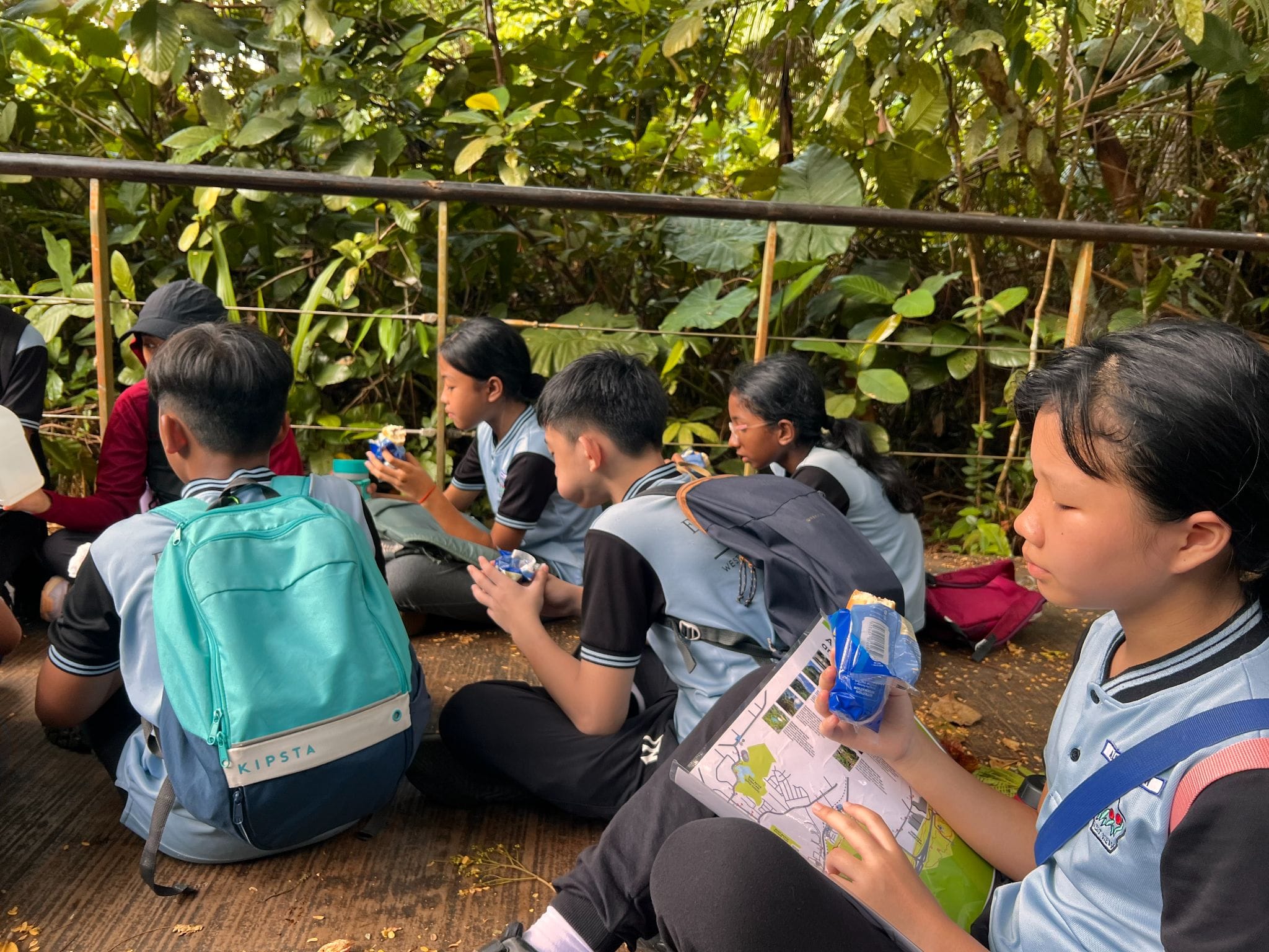 Students in blue uniforms sit on the ground eating snacks near a metal railing surrounded by dense green foliage.