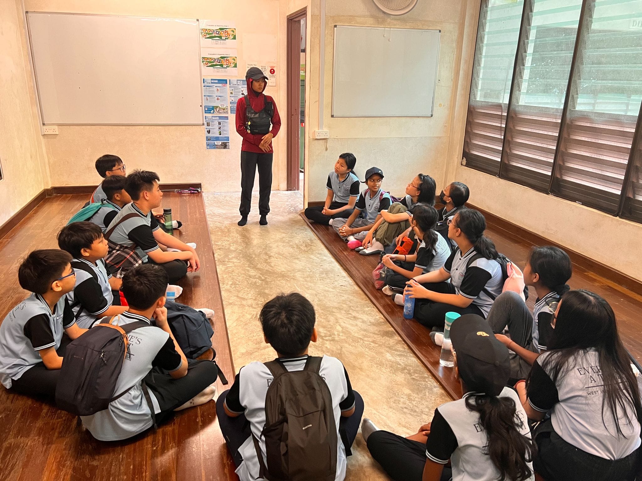 A standing adult in red addresses a group of uniformed students sitting on the floor in a classroom.
