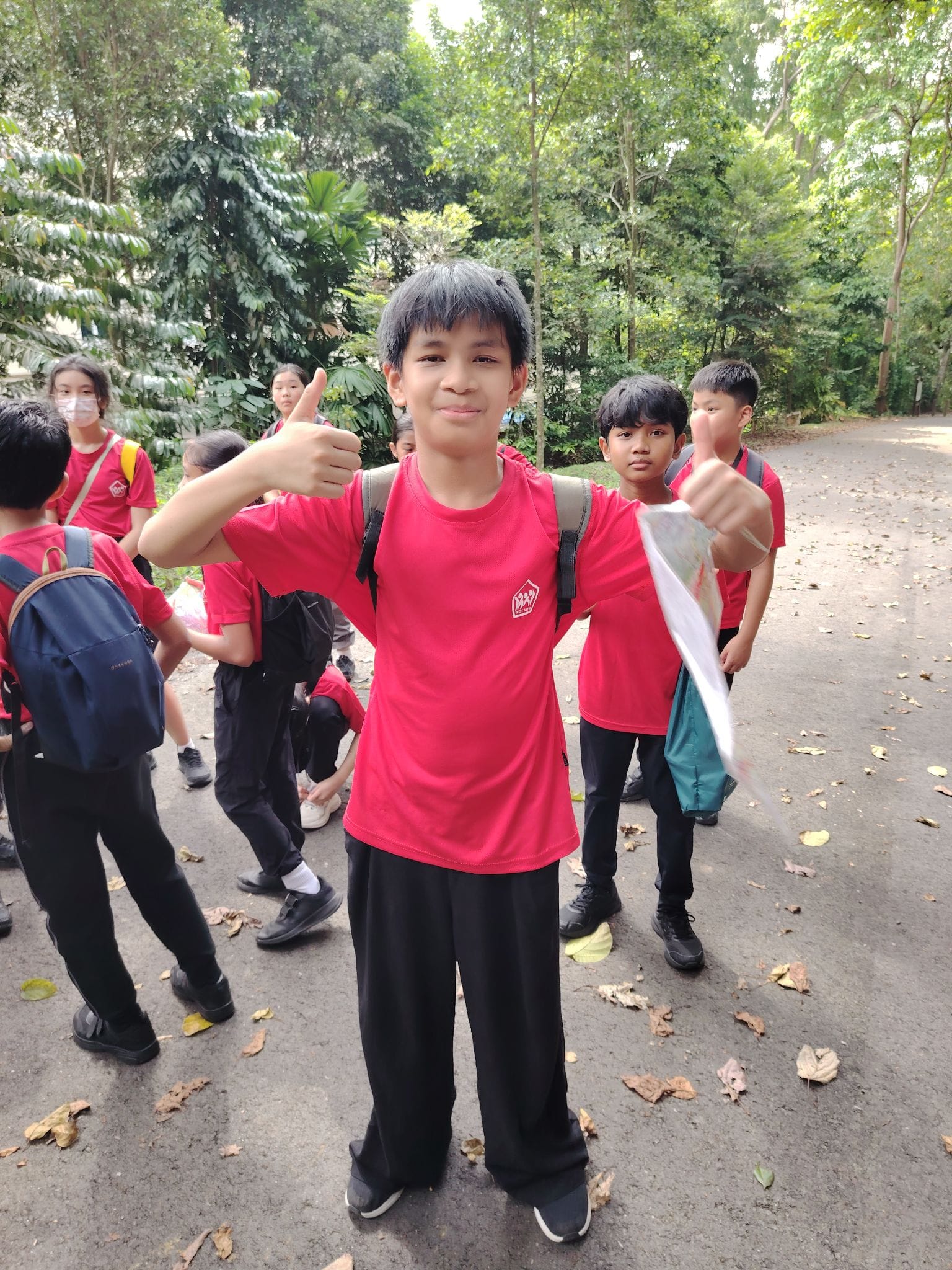 A smiling boy in a red shirt gives two thumbs up on a forest path, surrounded by classmates in matching uniforms.